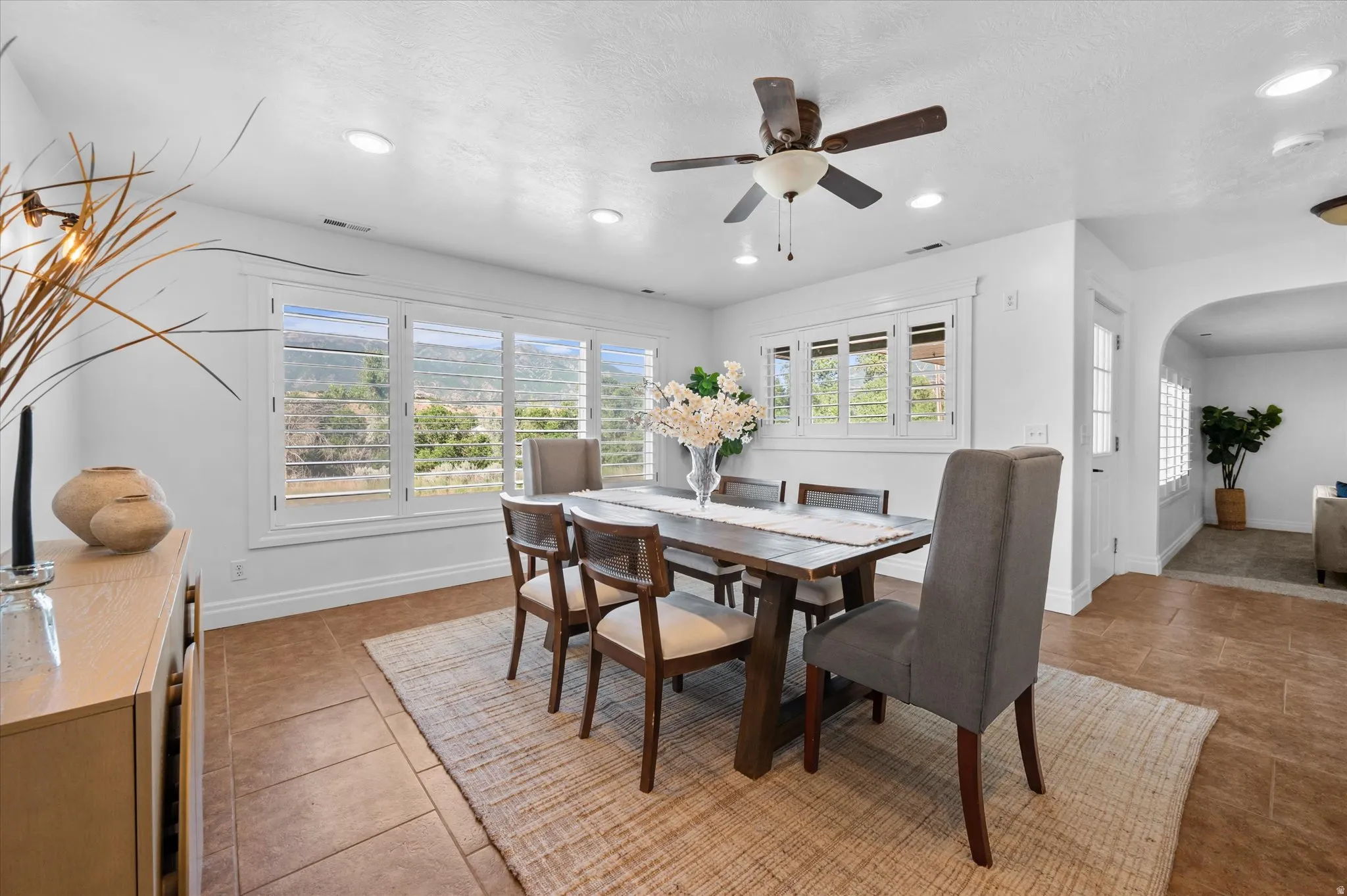 Dining area featuring large windows with white plantation shutters, a dark wood-finish dining table, and a ceiling fan with integrated lighting