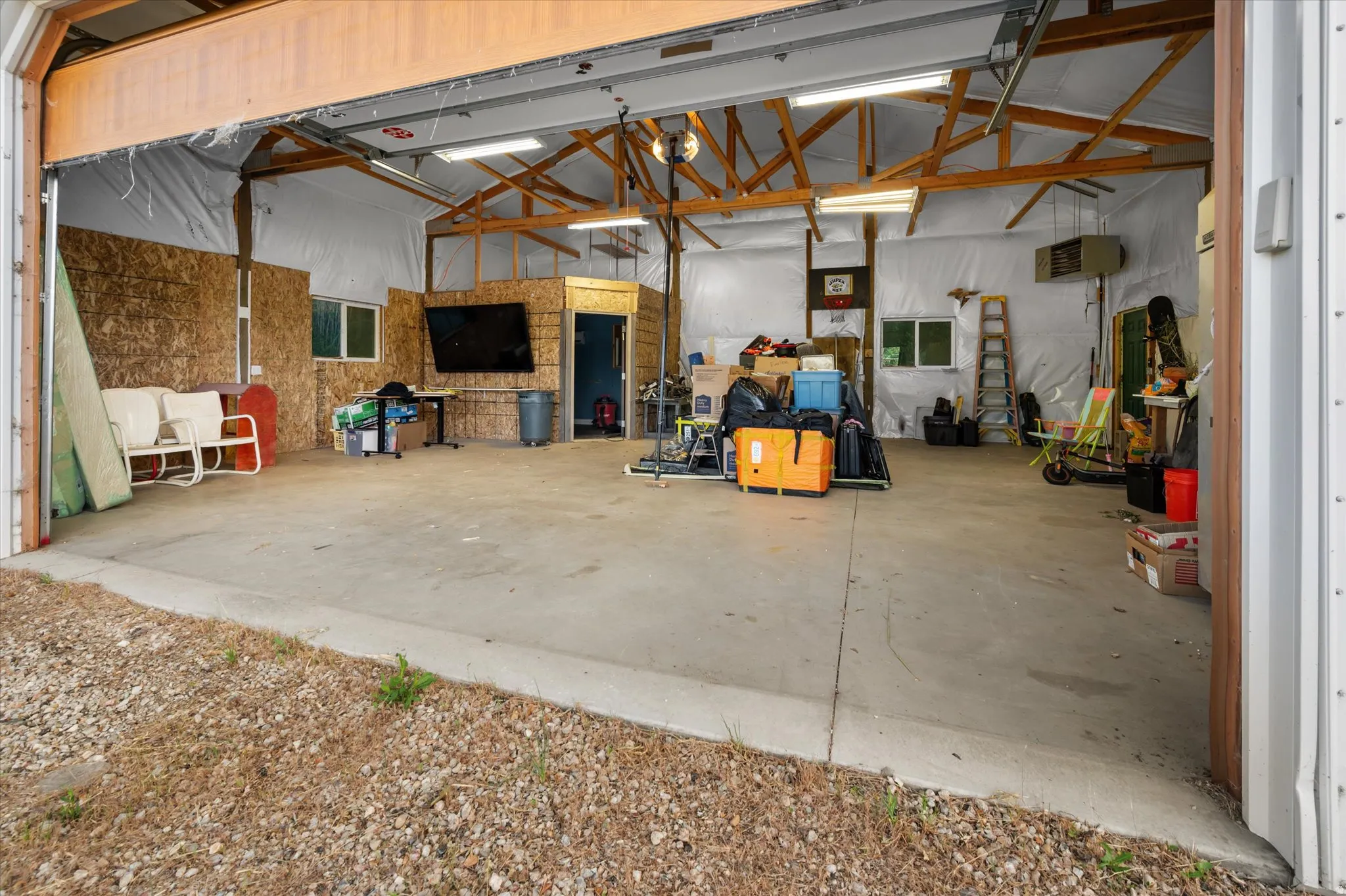 Spacious workshop featuring a large overhead door, concrete flooring, exposed wood beam ceiling, multiple windows, and fluorescent lighting