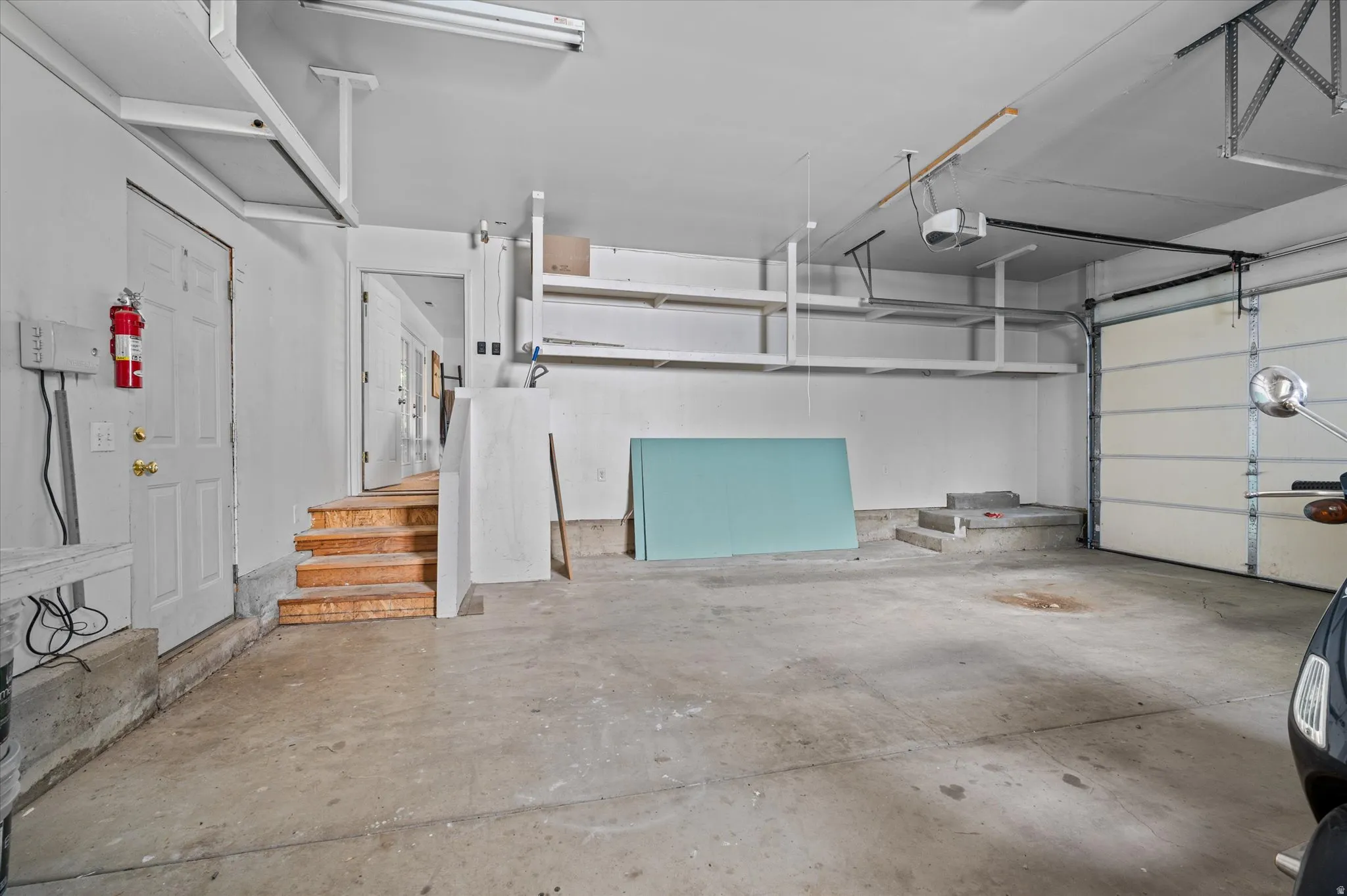 Spacious garage featuring concrete flooring, built-in overhead storage shelves, a full-panel garage door, and interior access via a paneled door with wooden stairs