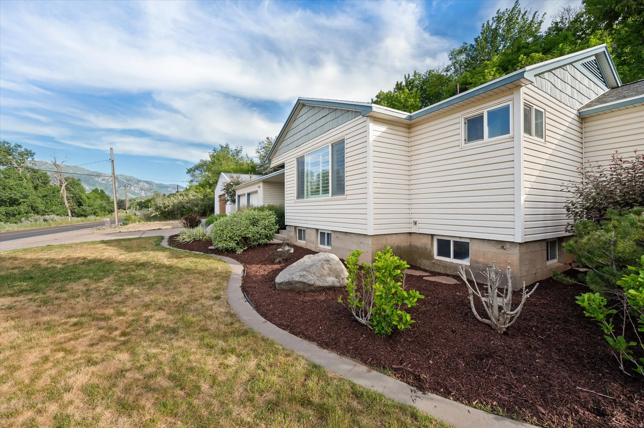 Cream-colored siding exterior with a light blue trim, featuring multiple windows and a landscaped bed with dark mulch and established plantings