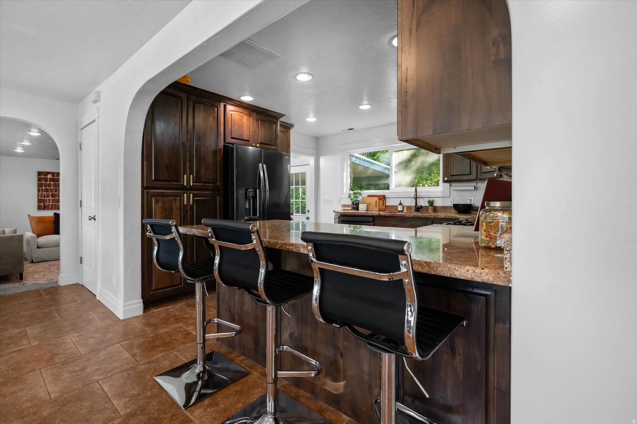 Kitchen island with stone-finish countertop and seating for three, featuring dark wood cabinetry, a black refrigerator, and a window above the sink