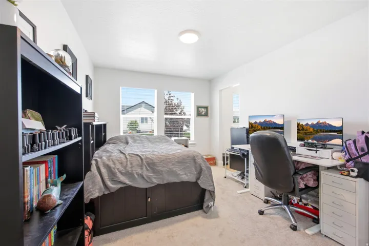Carpeted room with white walls, featuring a built-in dark wood bookshelf, a bed with integrated storage, and two windows with blinds