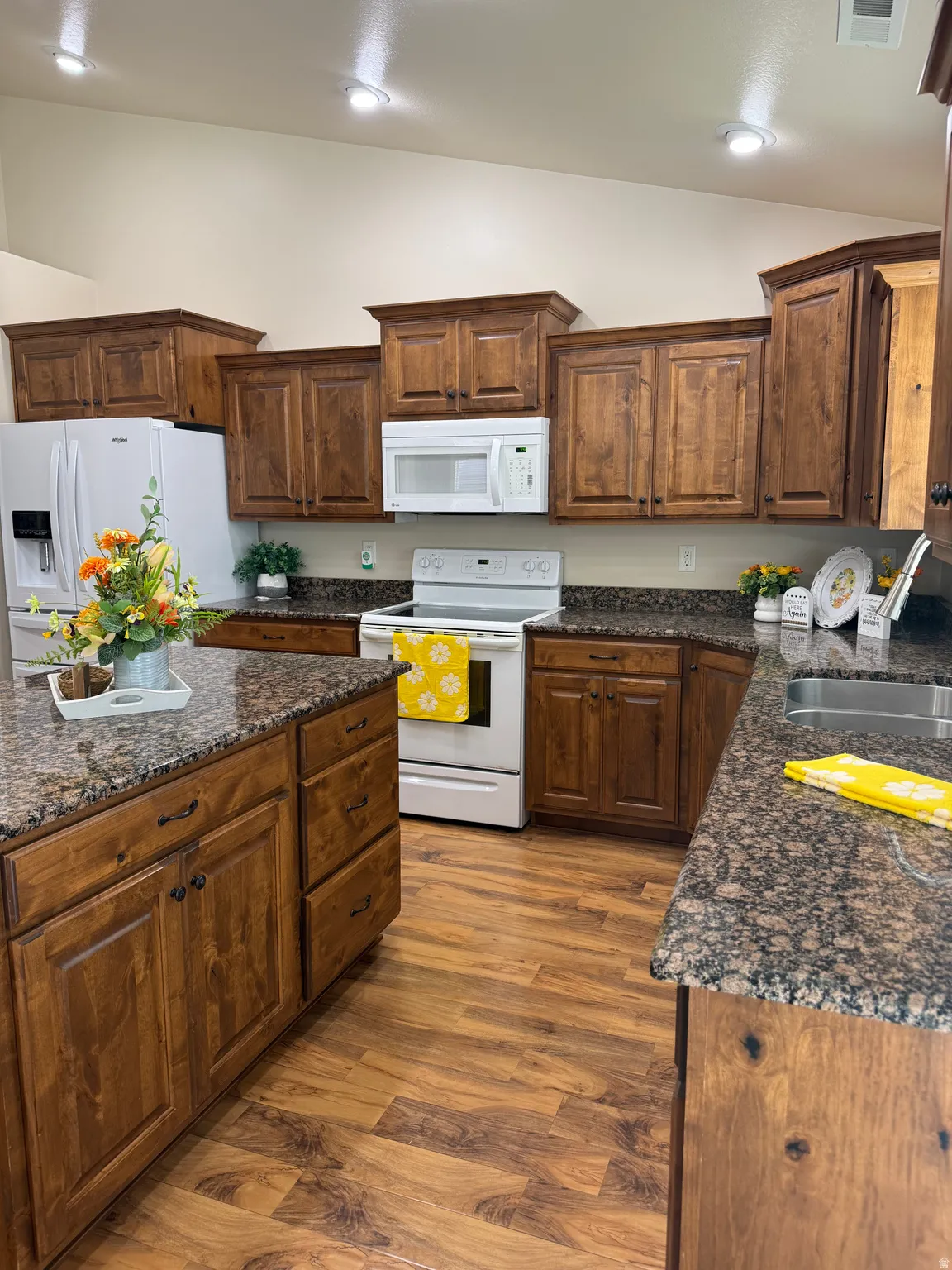 Kitchen featuring rich wood-finish cabinetry, granite-style countertops, wood-finish flooring, recessed lighting, and a vaulted ceiling