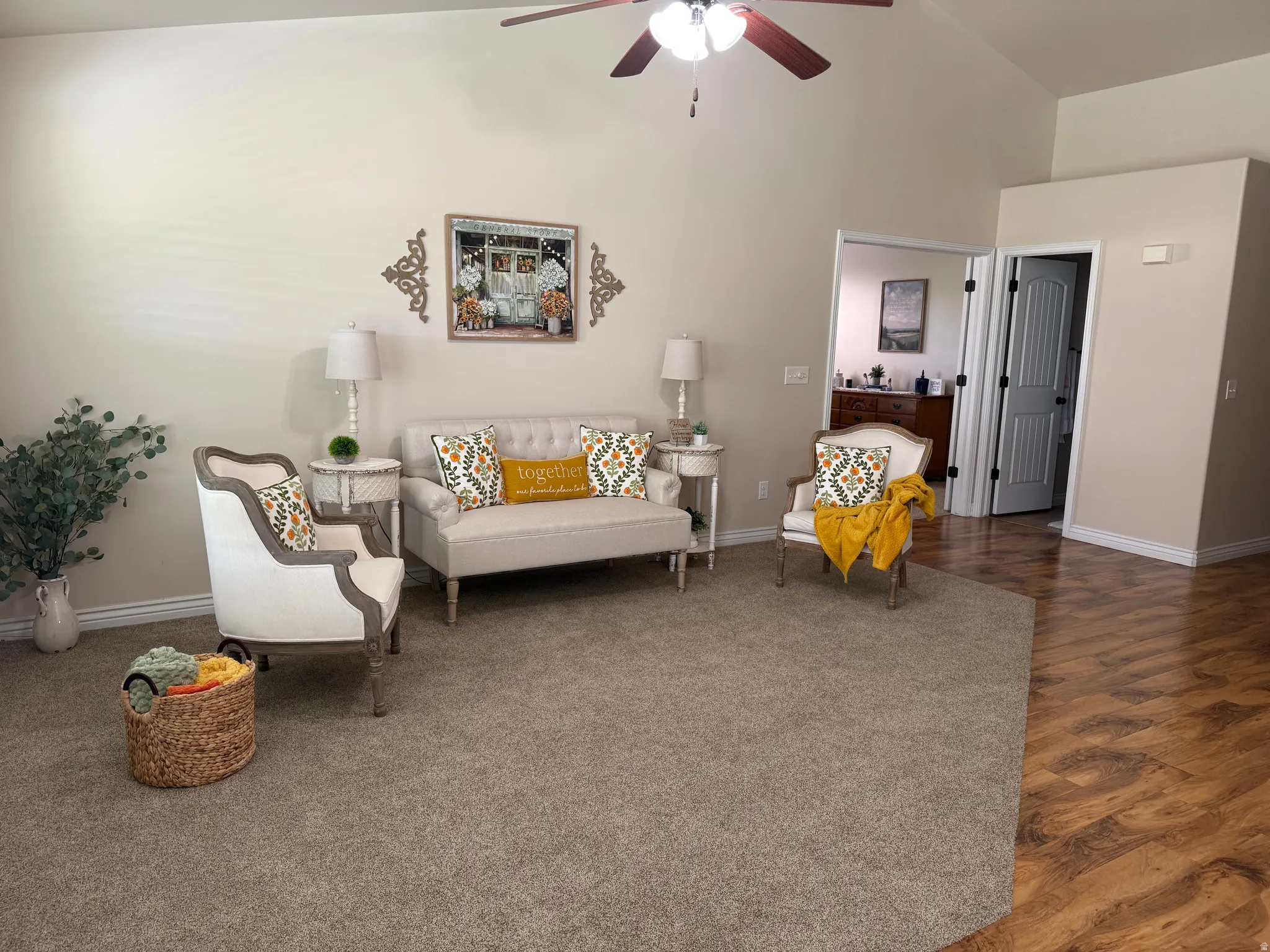 Vaulted ceiling living area featuring a ceiling fan, plush carpet, wood-finish flooring, and white baseboards