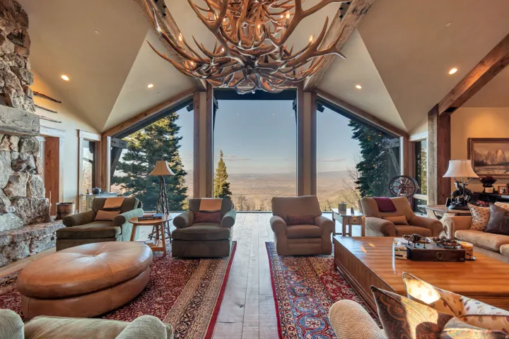 Vaulted living space featuring a large antler chandelier, exposed wood beams, a stone fireplace, and a wall of windows