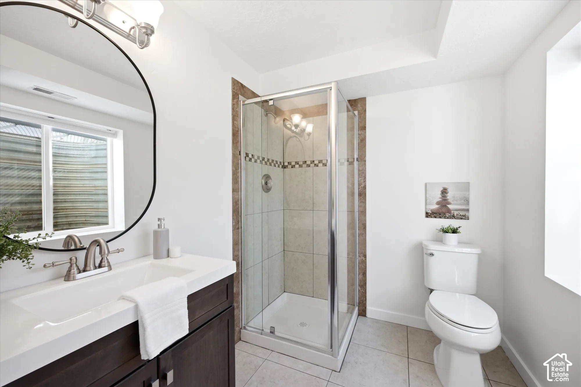 Contemporary bathroom featuring a dark wood vanity with a white countertop, an oval-framed mirror, and a glass-enclosed shower with tiled walls and a decorative accent band