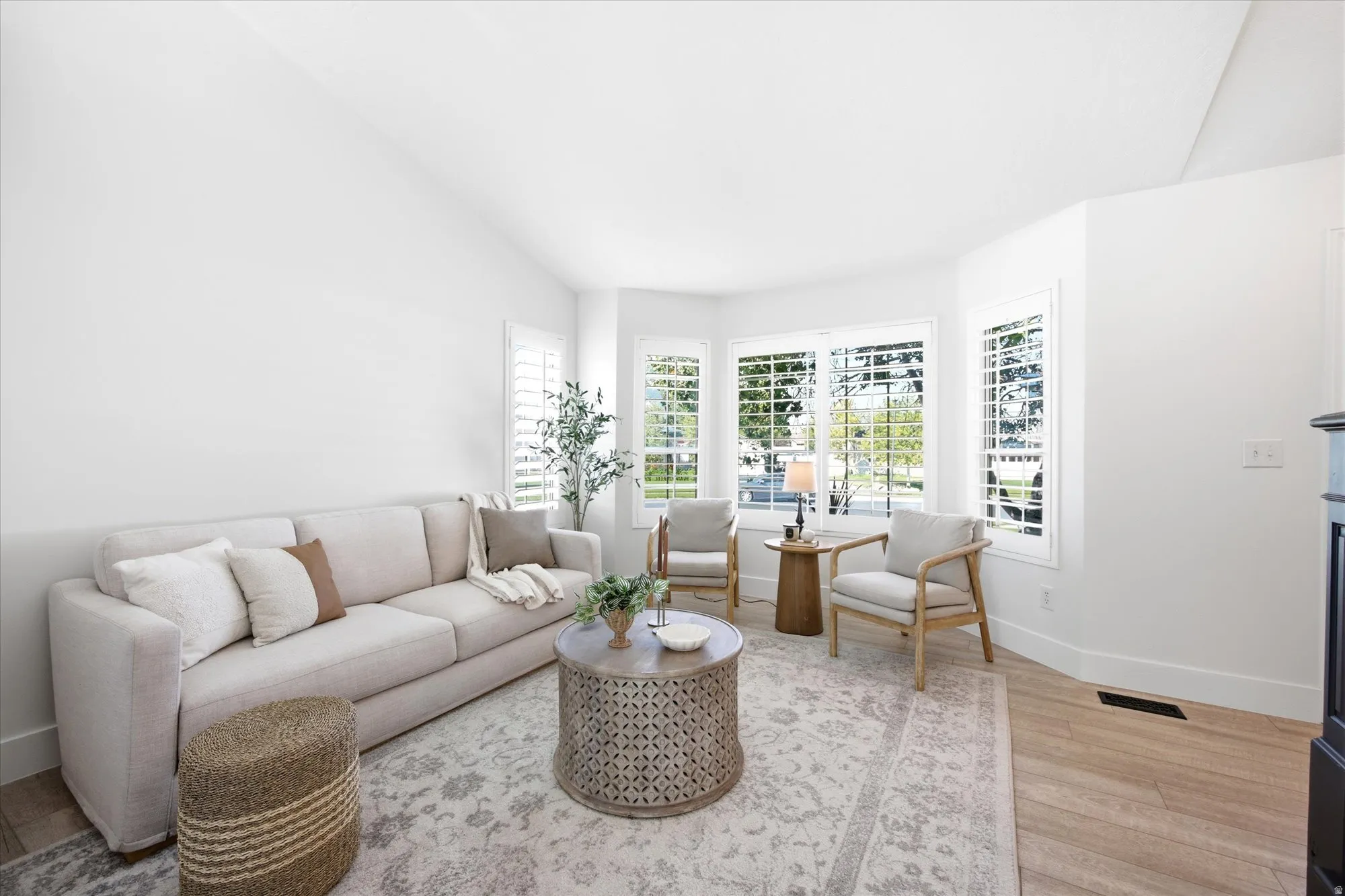 Vaulted ceiling living area featuring a bay window with plantation shutters, wood-finish flooring, and a fireplace with a dark mantel
