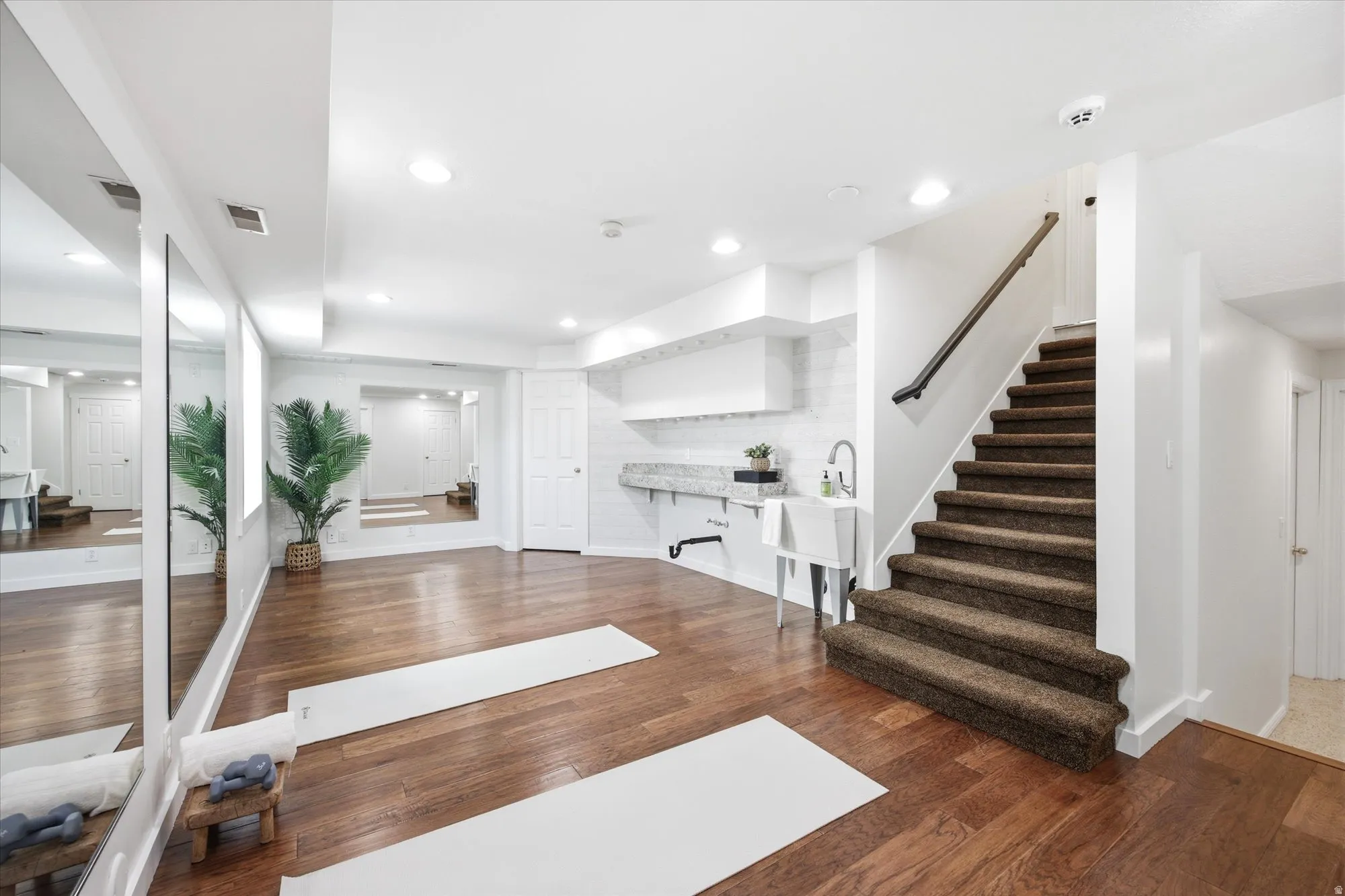 Spacious lower-level area with wood-finish flooring, a built-in wet bar featuring a utility sink and stone countertop, and full-wall mirrors