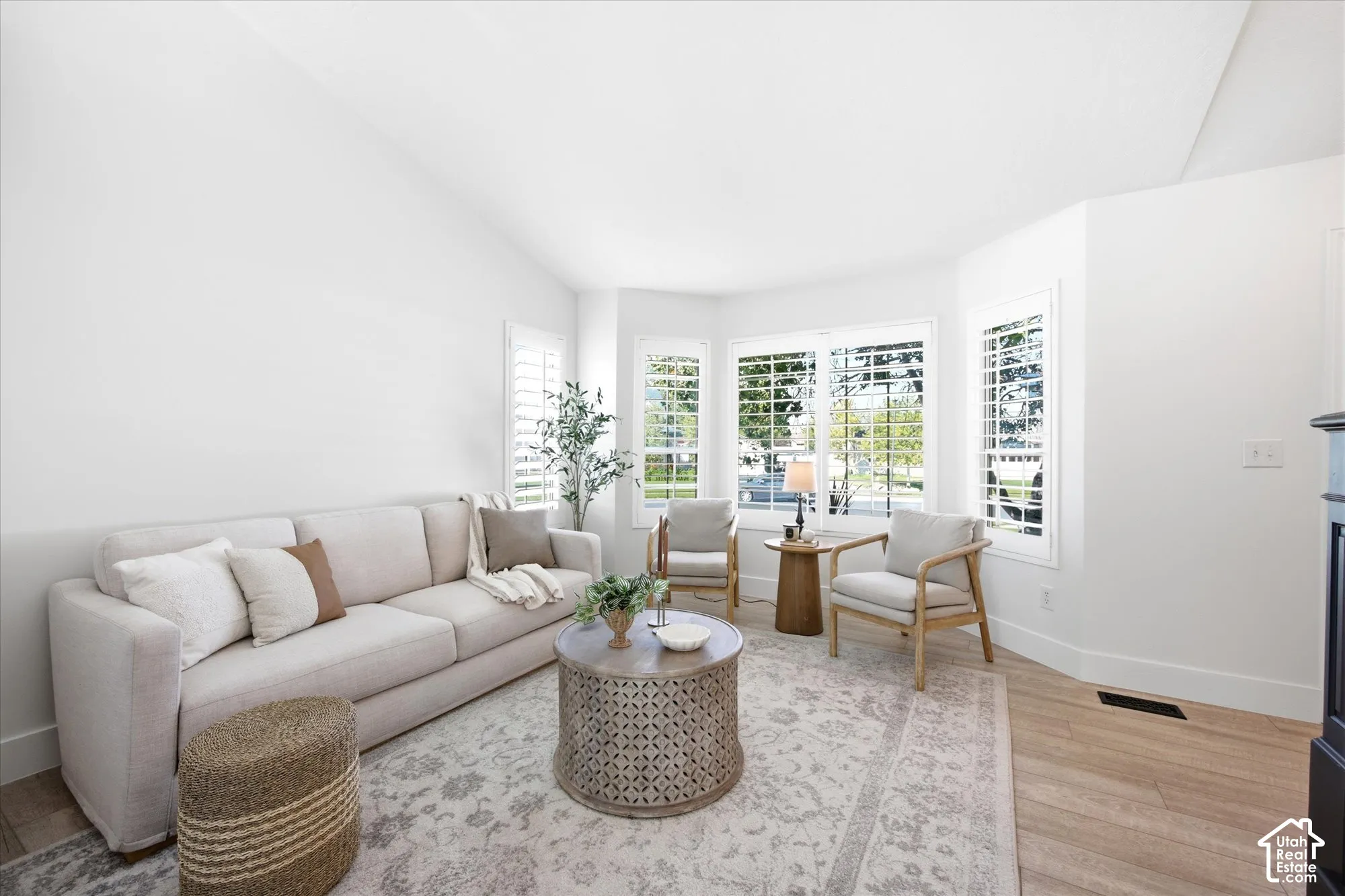 Vaulted ceiling living area featuring a bay window with plantation shutters, wood-finish flooring, and a fireplace with a dark mantel