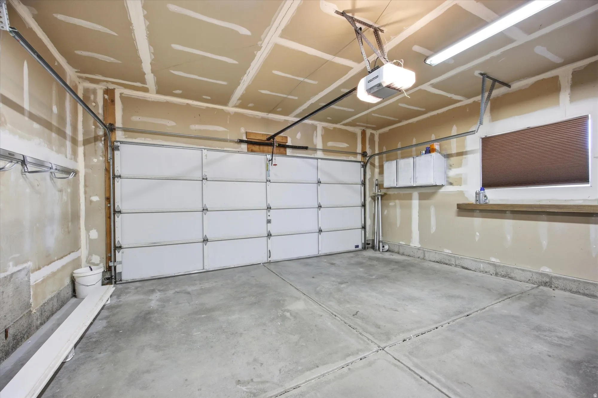 Concrete flooring garage featuring a sectional overhead door with an automatic opener, a window with a mounted blind, and built-in overhead fluorescent lighting