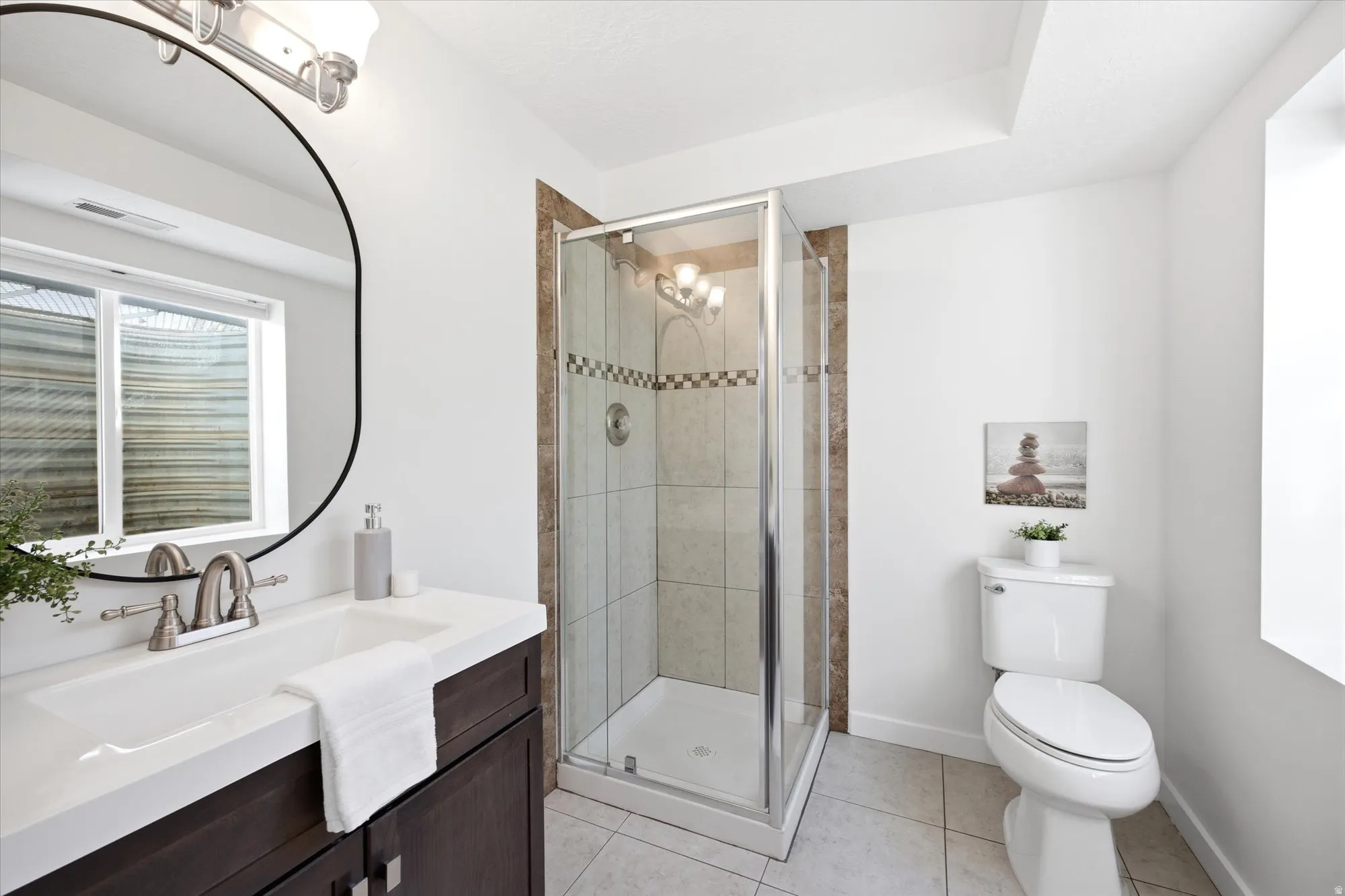 Contemporary bathroom featuring a dark wood vanity with a white countertop, an oval-framed mirror, and a glass-enclosed shower with tiled walls and a decorative accent band