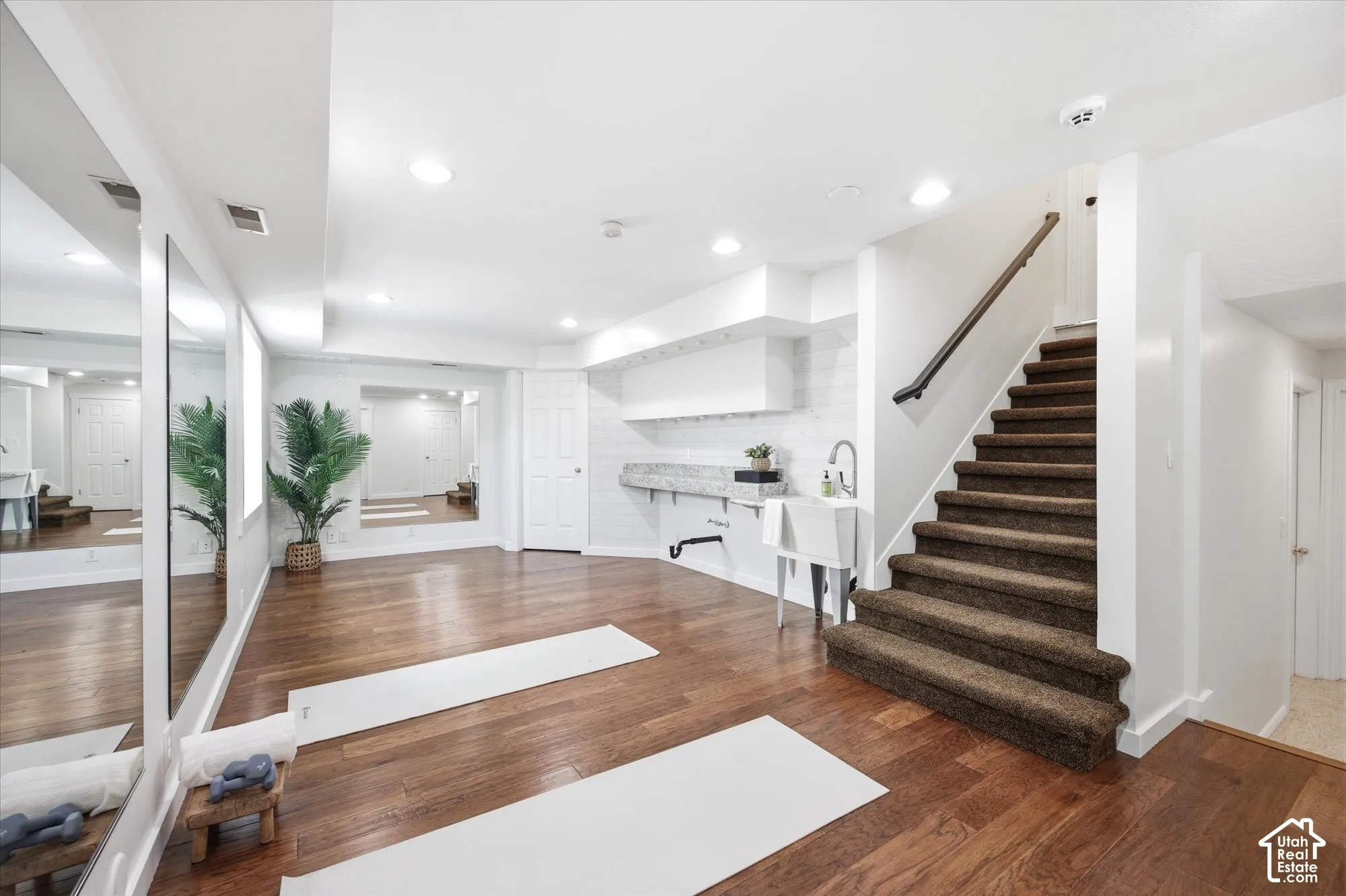 Spacious lower-level area with wood-finish flooring, a built-in wet bar featuring a utility sink and stone countertop, and full-wall mirrors