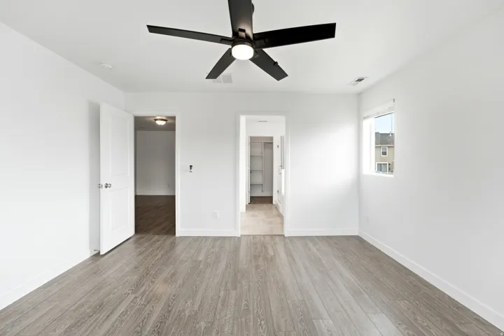 Spacious room featuring wood-finish flooring, a modern black ceiling fan with integrated lighting, white walls, and a window with a roller shade