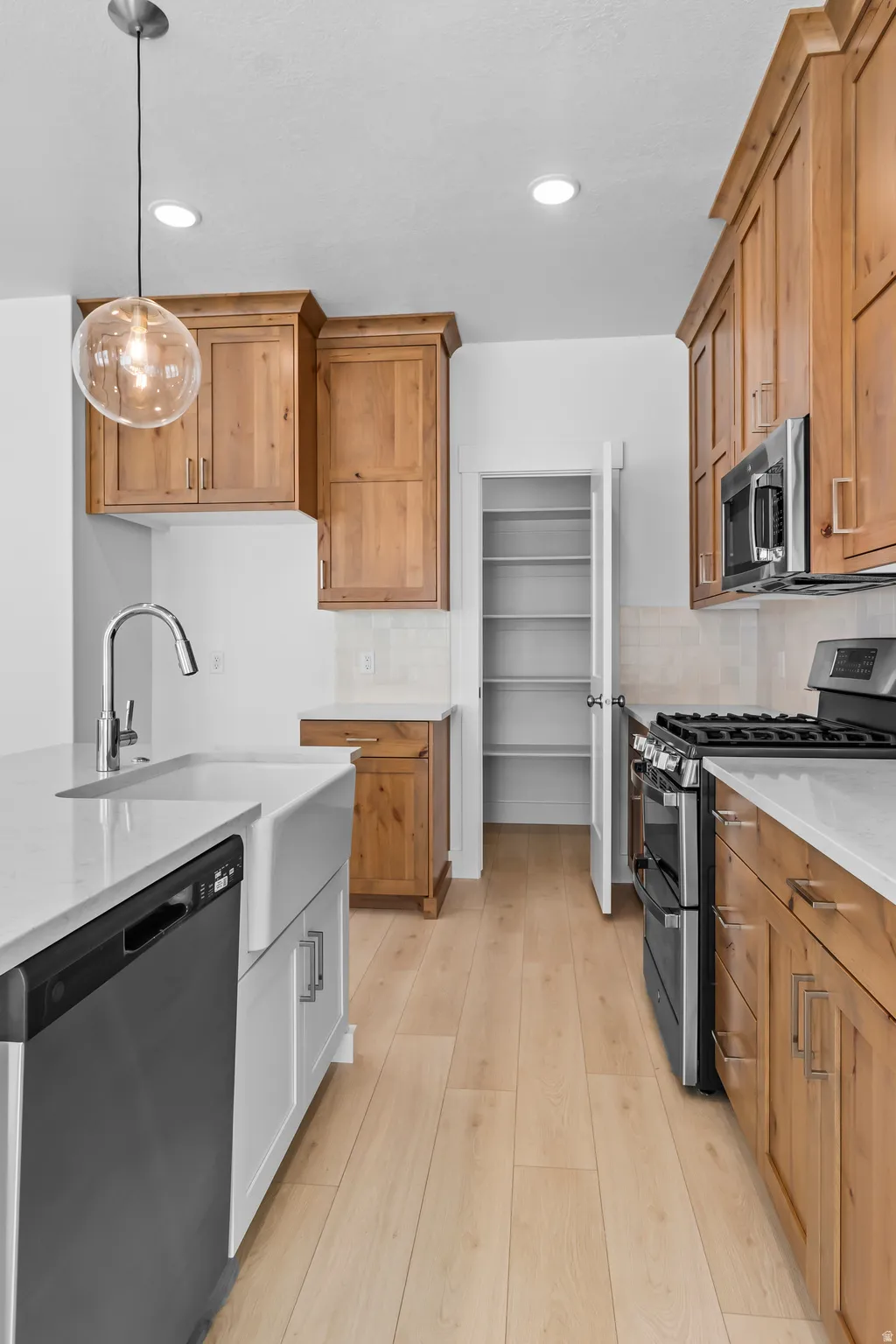 Modern kitchen featuring wood-finish flooring, white perimeter cabinetry with a farmhouse-style sink, stainless steel appliances, and a pantry with built-in shelving