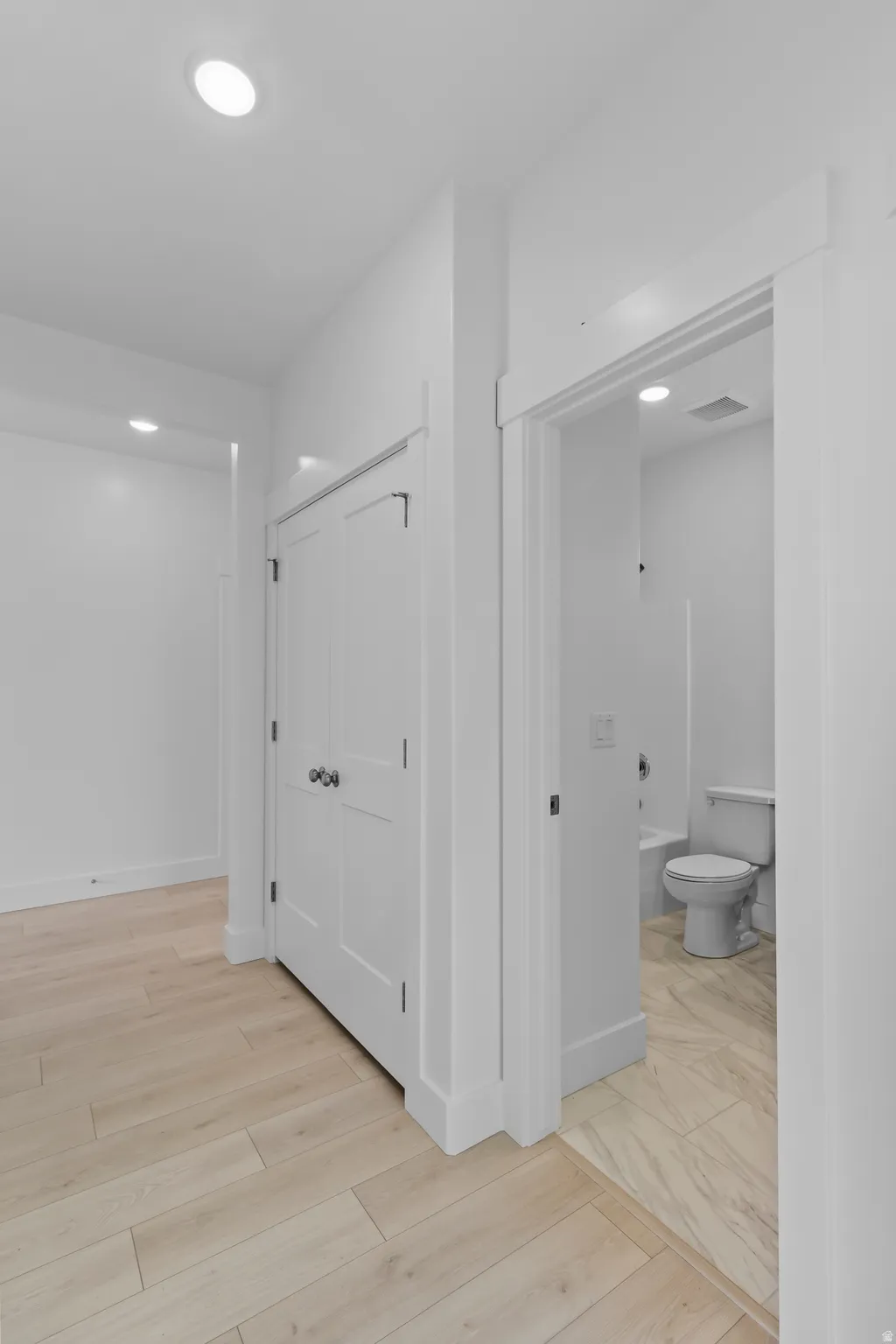Light wood-finish flooring extending into a white-trimmed doorway, revealing a bathroom with a white toilet and tub