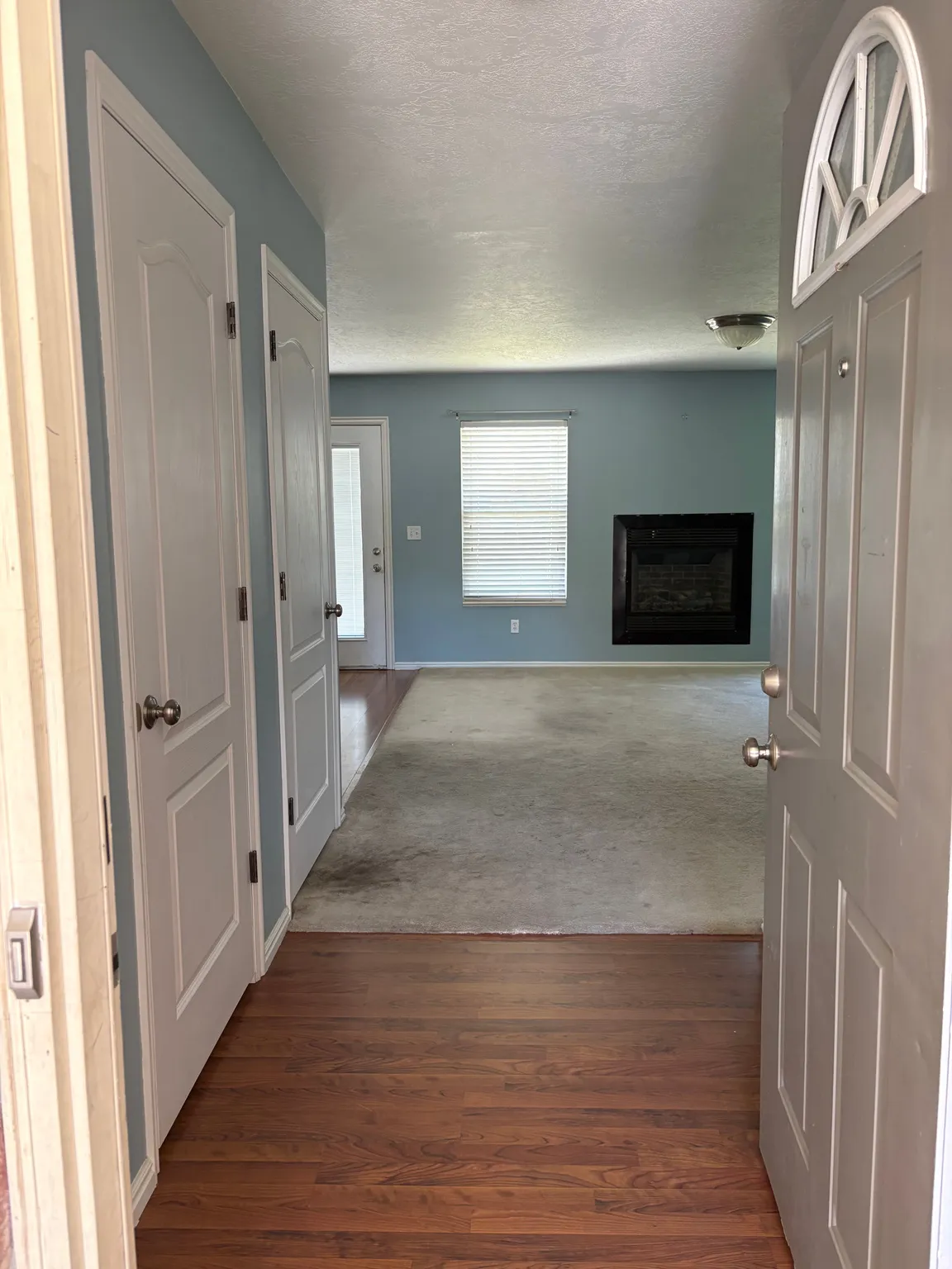 Entryway featuring wood-finish flooring, leading to a living area with light blue walls, a fireplace with a dark surround, and a window with blinds