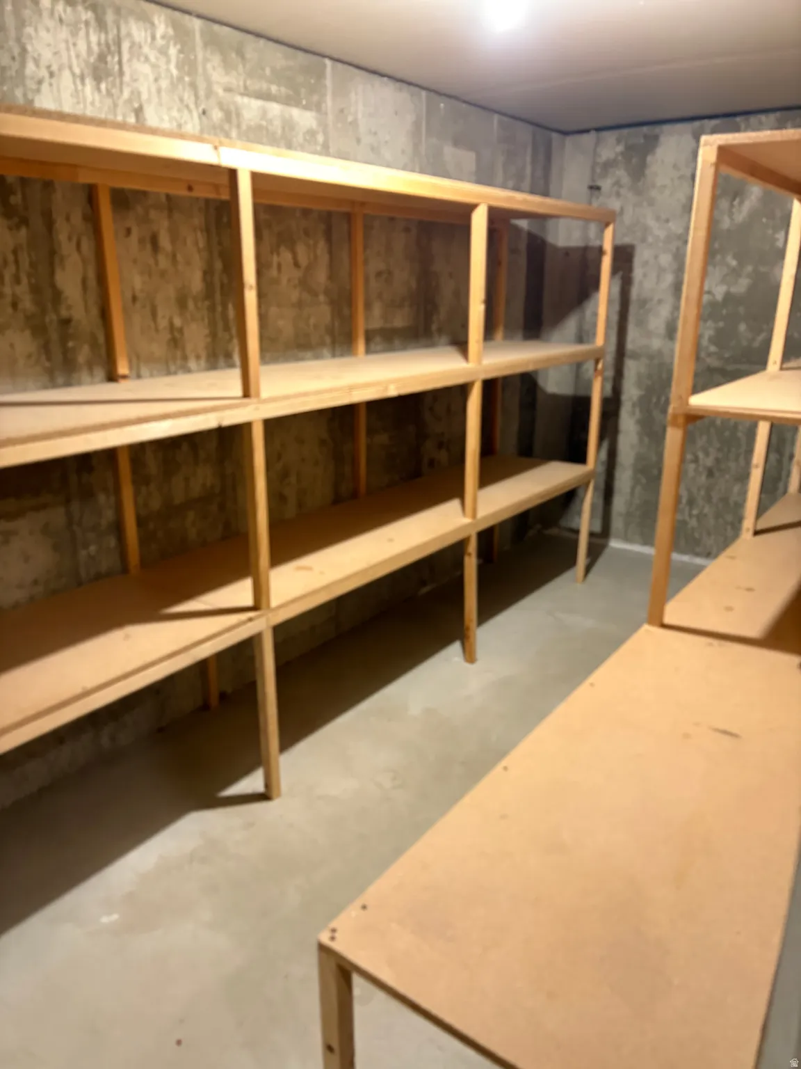 Utility area featuring extensive built-in wooden shelving units, concrete walls, concrete flooring, and overhead lighting