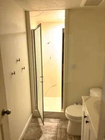 Bathroom featuring a glass-enclosed shower, a white vanity with an integrated sink, dark cabinet hardware, a toilet, and neutral-toned tiled flooring