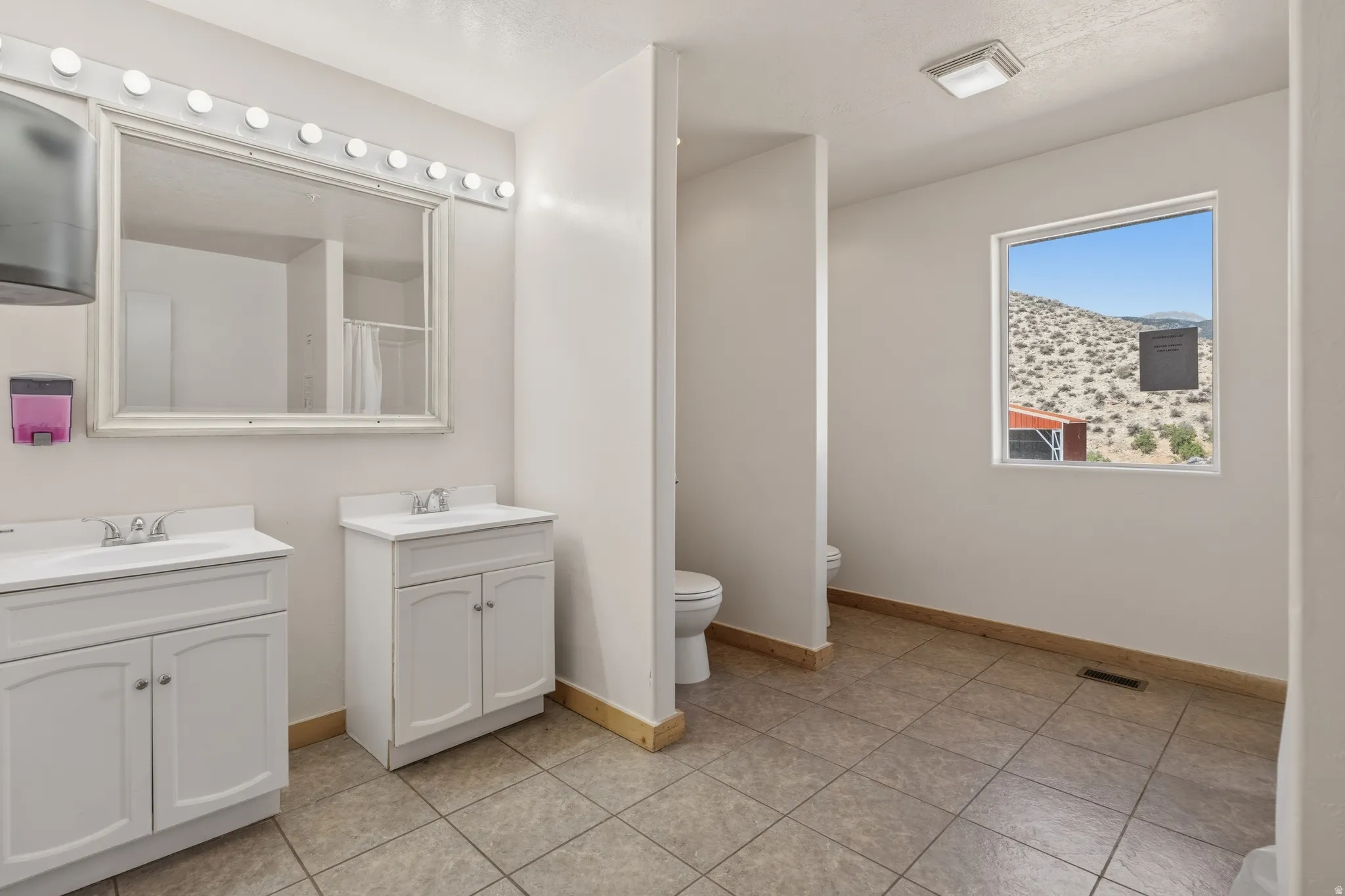 Spacious restroom featuring dual white vanity cabinets, a large mirror with strip lighting, and a square window offering desert and mountain scenery