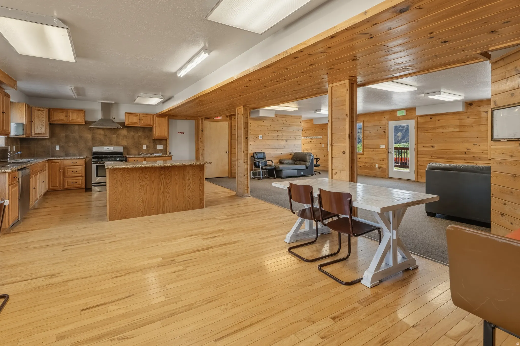 Open-concept kitchen and dining area featuring wood-finish flooring, wood paneling, granite-style countertops, stainless steel appliances, and an island with bar seating