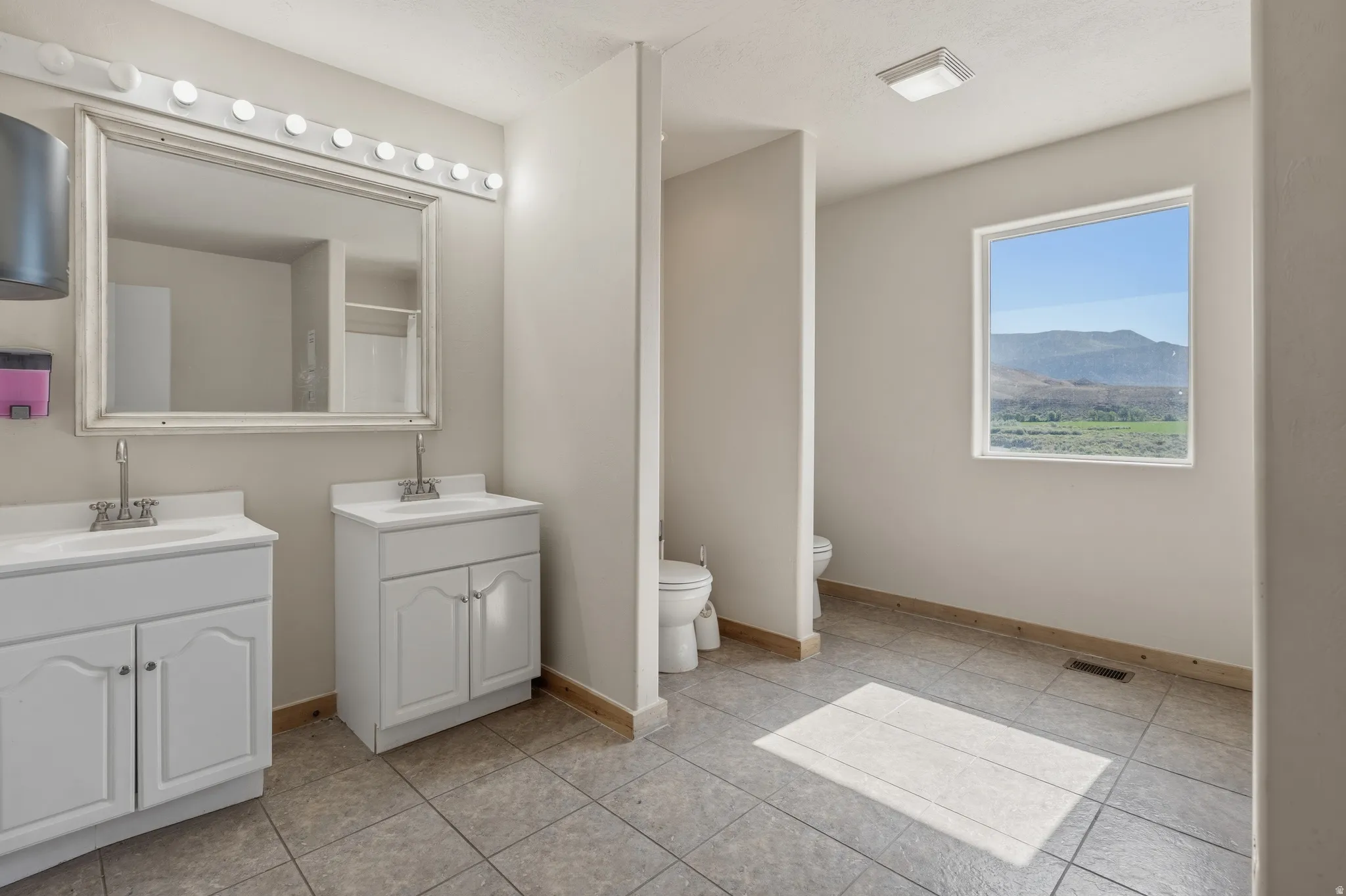 Spacious bathroom featuring dual white vanity cabinets with integrated sinks, a large framed mirror with Hollywood lighting, and light grey floor tiling