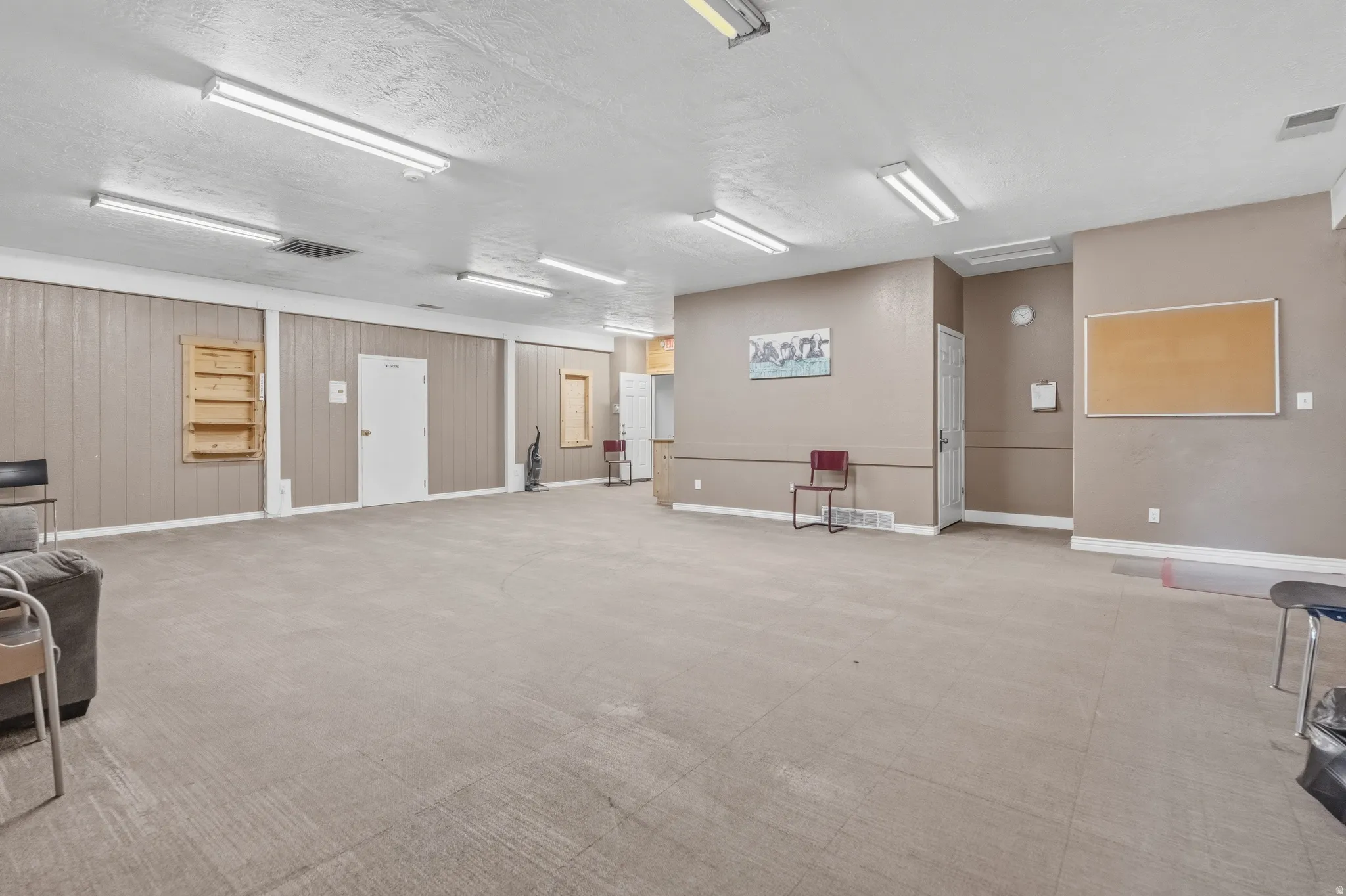 Expansive open space featuring light-colored flooring, neutral wall tones, multiple overhead fluorescent lighting fixtures, and a white interior door
