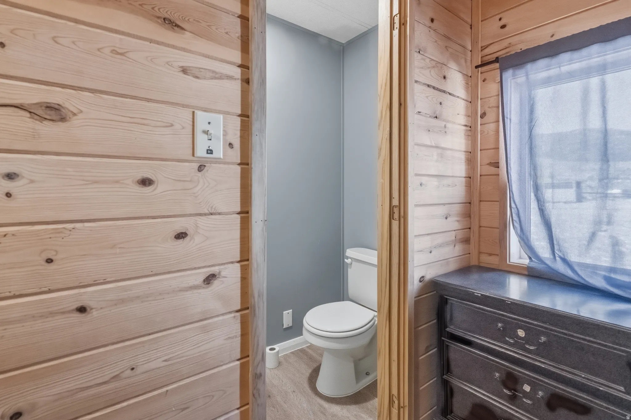 Wood-paneled wall with a light switch, opening to a powder room featuring a white toilet and light wood-finish flooring
