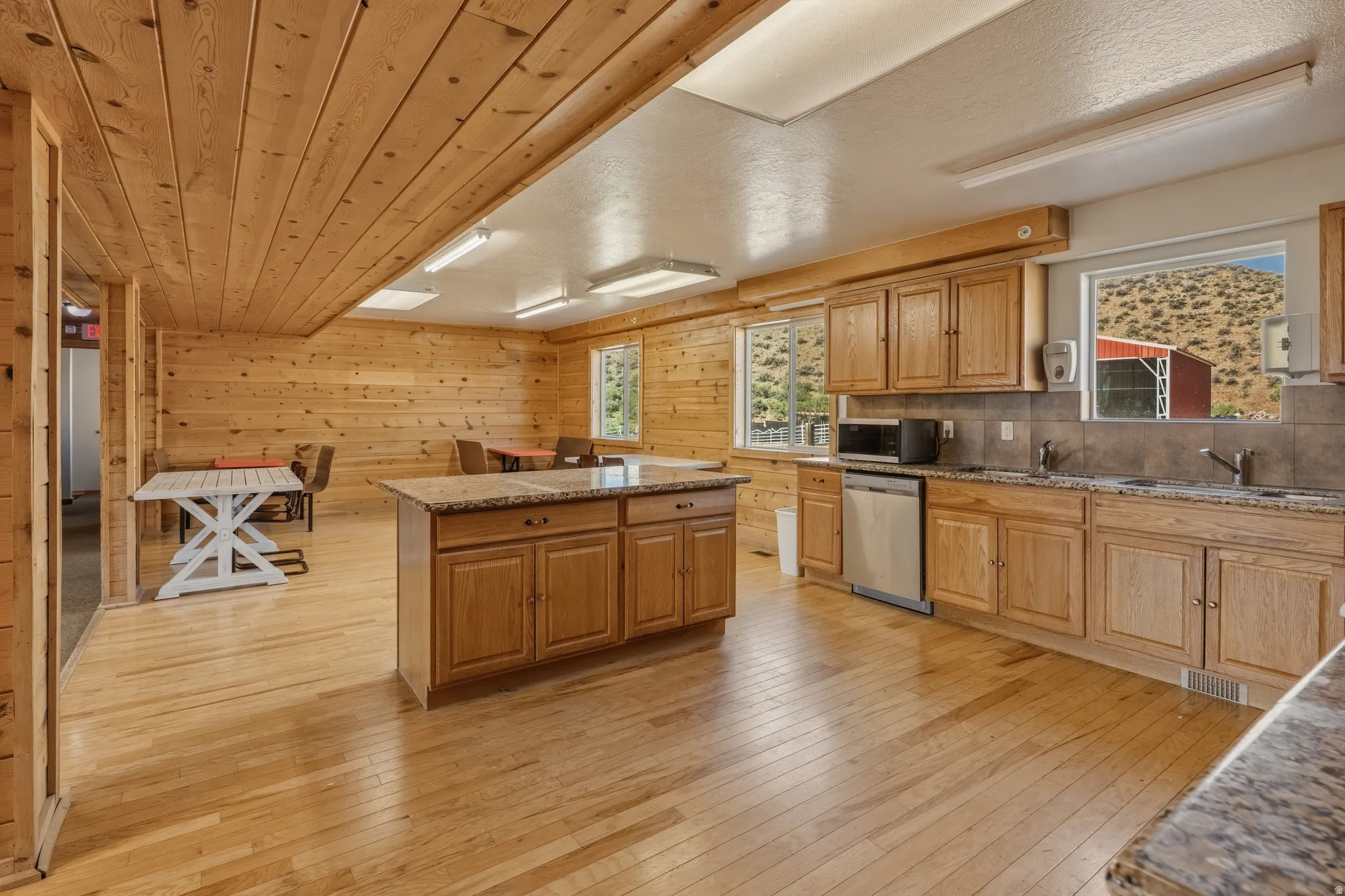 Spacious kitchen and dining area featuring natural wood paneling, light wood-finish flooring, a central island with a stone countertop, wood cabinetry, and stainless steel appliances