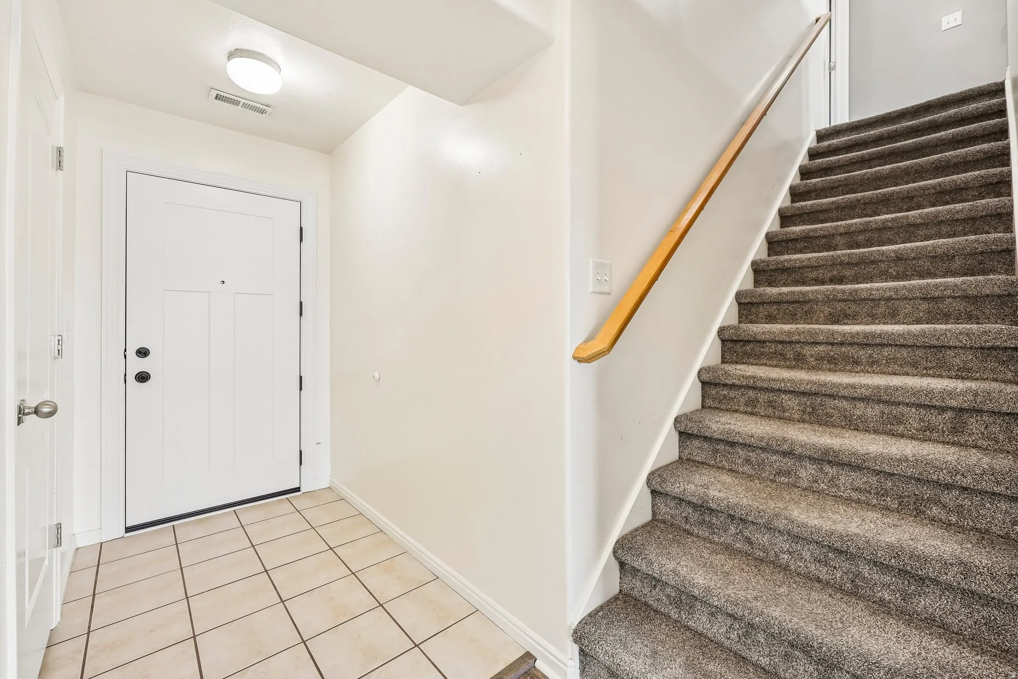 Entryway featuring a white paneled door with dark hardware, square tile flooring, and a carpeted staircase with a wood handrail