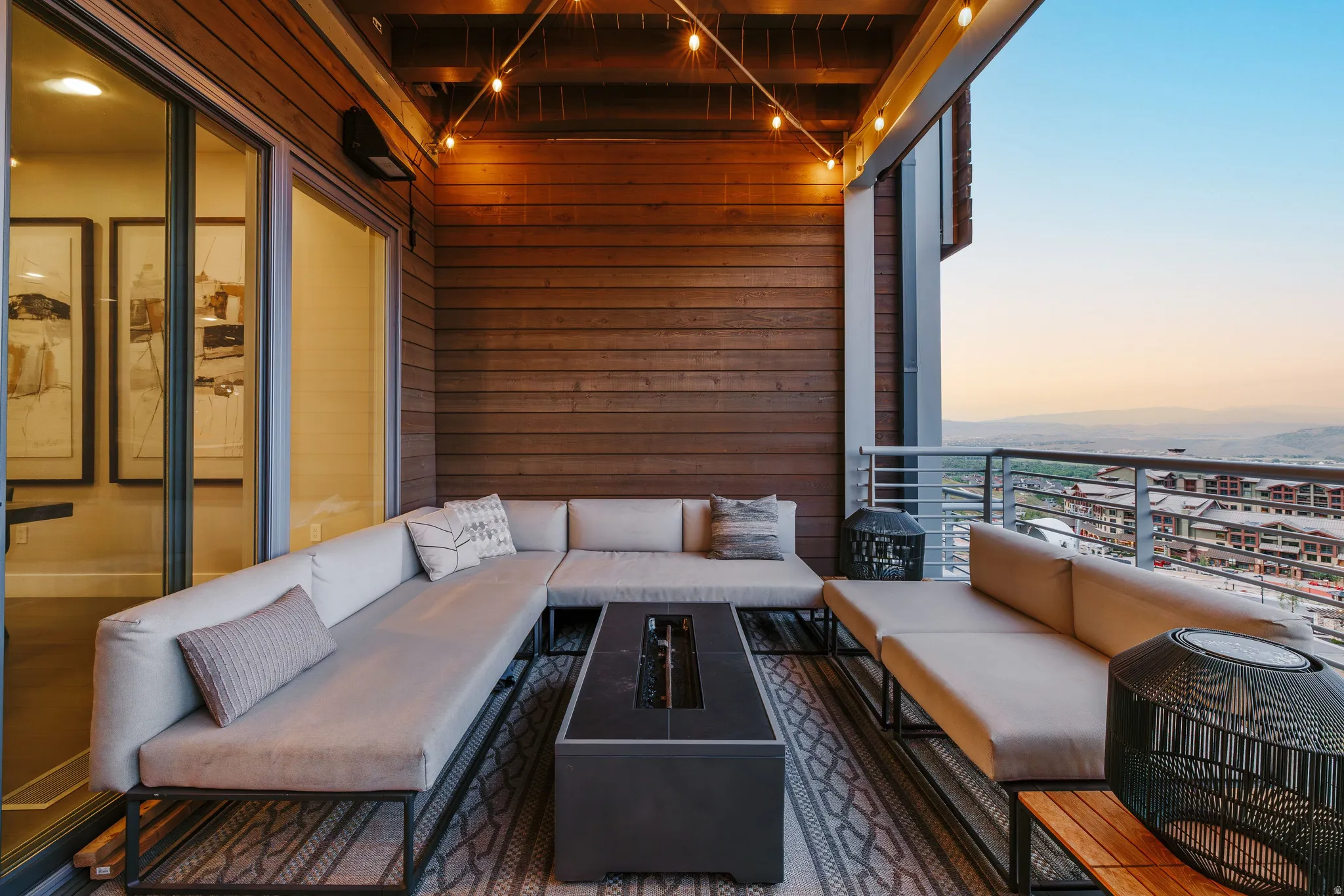 Spacious outdoor balcony with wood-paneled walls and ceiling, featuring a built-in fire pit, string lighting, and a metal railing