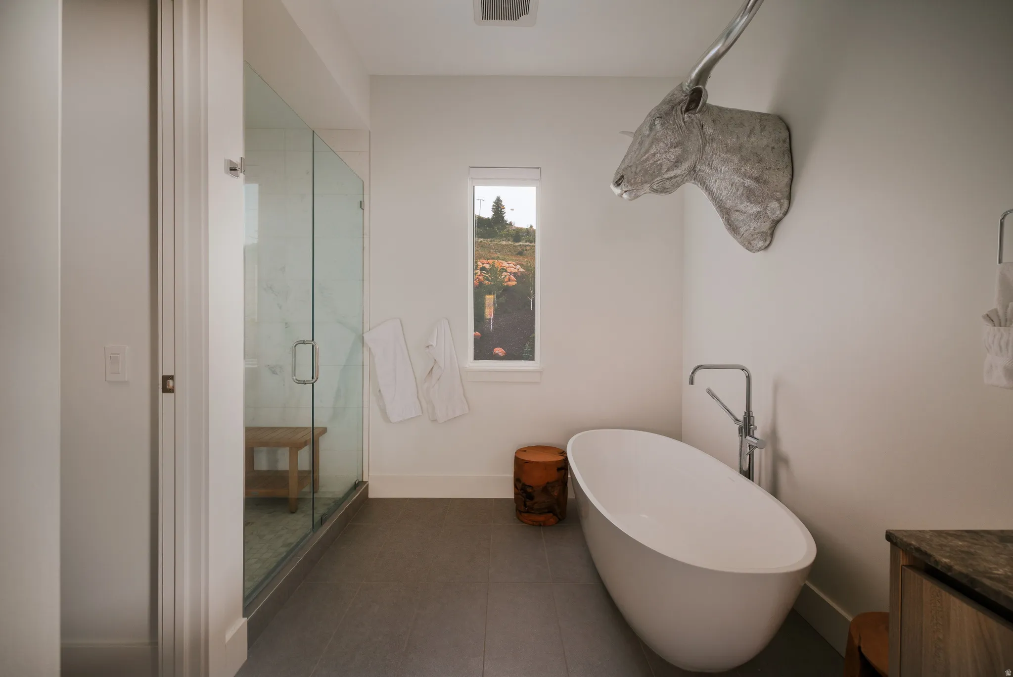 Freestanding soaking tub with floor-mounted faucet, glass-enclosed shower, dark floor tiling, and a single window