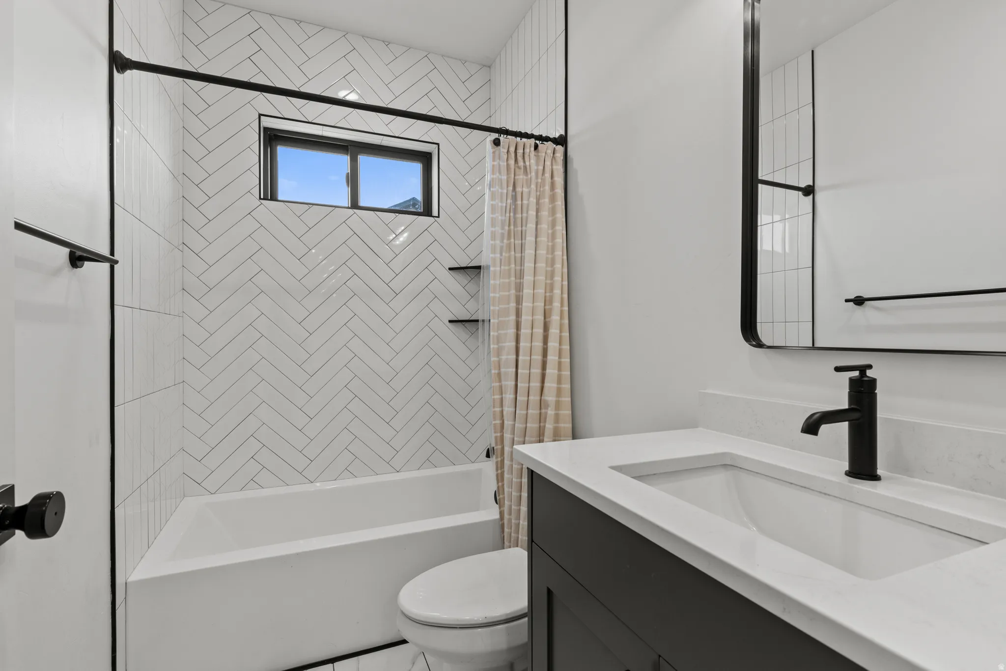 Bathroom featuring a white subway tile shower surround in a herringbone pattern with built-in shelving, a single vanity with a white countertop and integrated sink, a rectangular mirror with a black frame, and matte black fixtures throughout