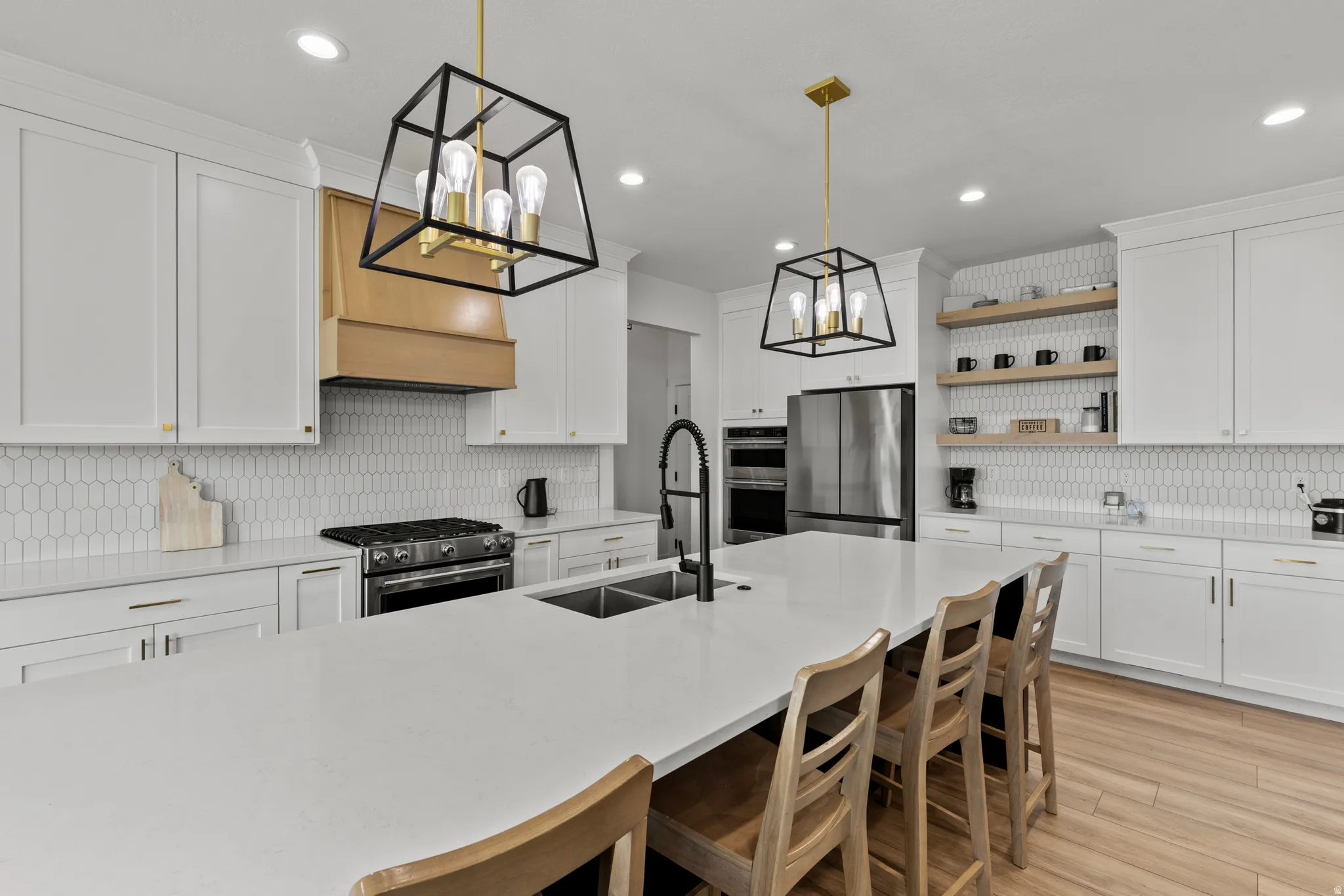 Spacious kitchen featuring an expansive central island with integrated double sink, white cabinetry, a large range with a wood-finish vent hood, a full-size stainless steel refrigerator, and wood-finish flooring