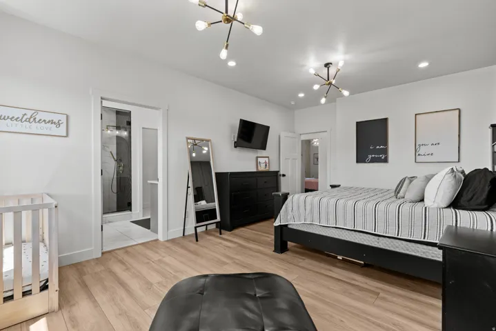 Spacious bedroom featuring wood-finish flooring, two modern sputnik chandeliers, recessed lighting, and a private en-suite bathroom with a glass-enclosed shower