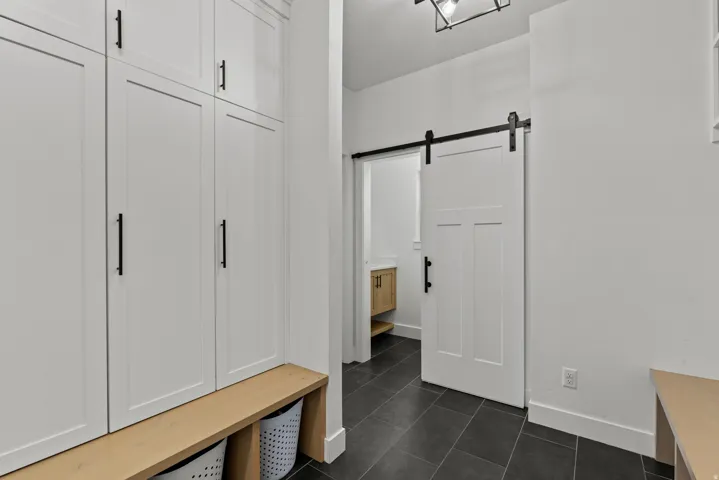 White shaker-style cabinetry with black hardware, natural wood-finish bench, dark tile flooring, and a sliding barn door with black track