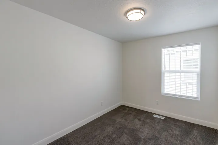 Neutral-toned room featuring a single window with blinds, a flush-mount ceiling light fixture, plush dark gray carpeting, and white baseboards