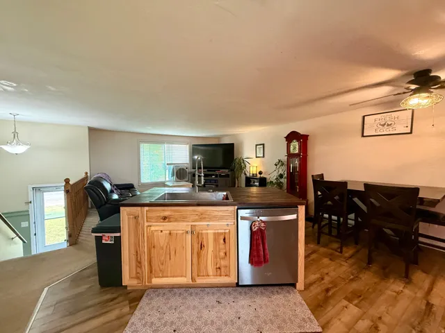 Open-concept kitchen and living area featuring wood-finish flooring, a central island with natural wood cabinetry and an integrated sink, a stainless steel dishwasher, and a dining area with a ceiling fan light fixture