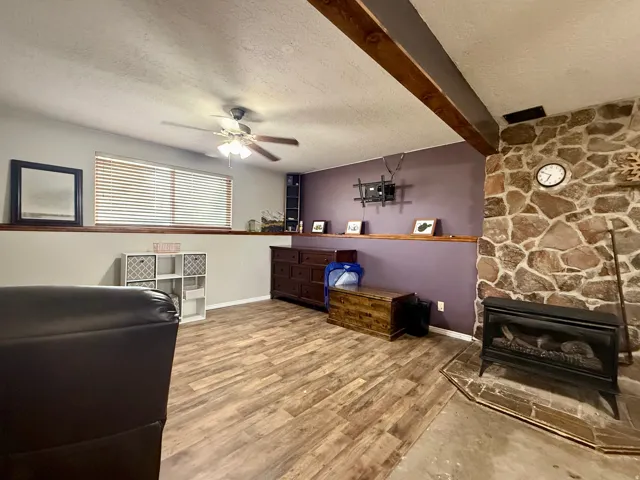 Recreation room featuring a stone-clad fireplace with hearth, wood-finish flooring, a ceiling fan, and exposed wood ceiling beams