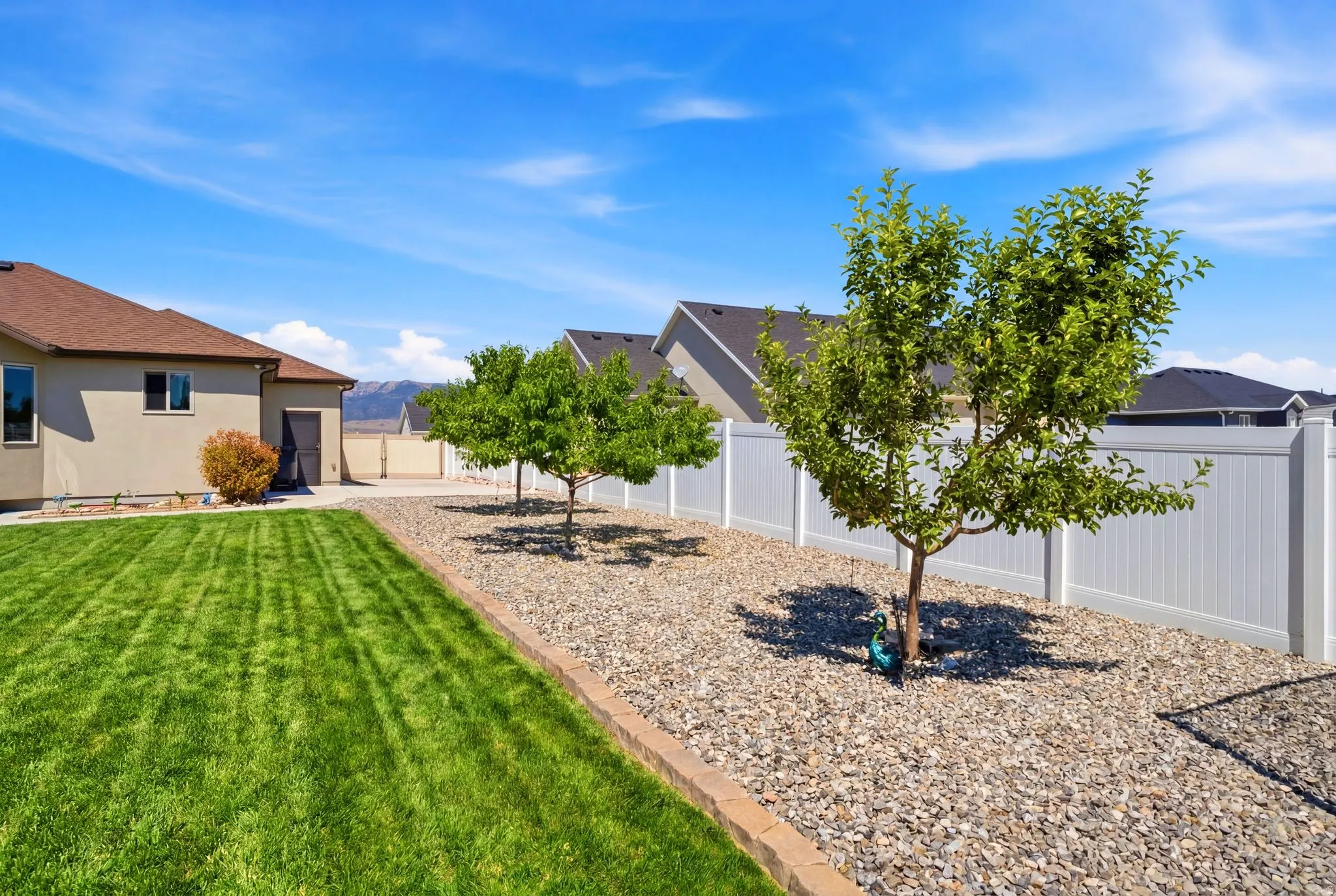 Expansive backyard featuring a manicured lawn, established trees, a stone-lined garden bed, and a white privacy fence