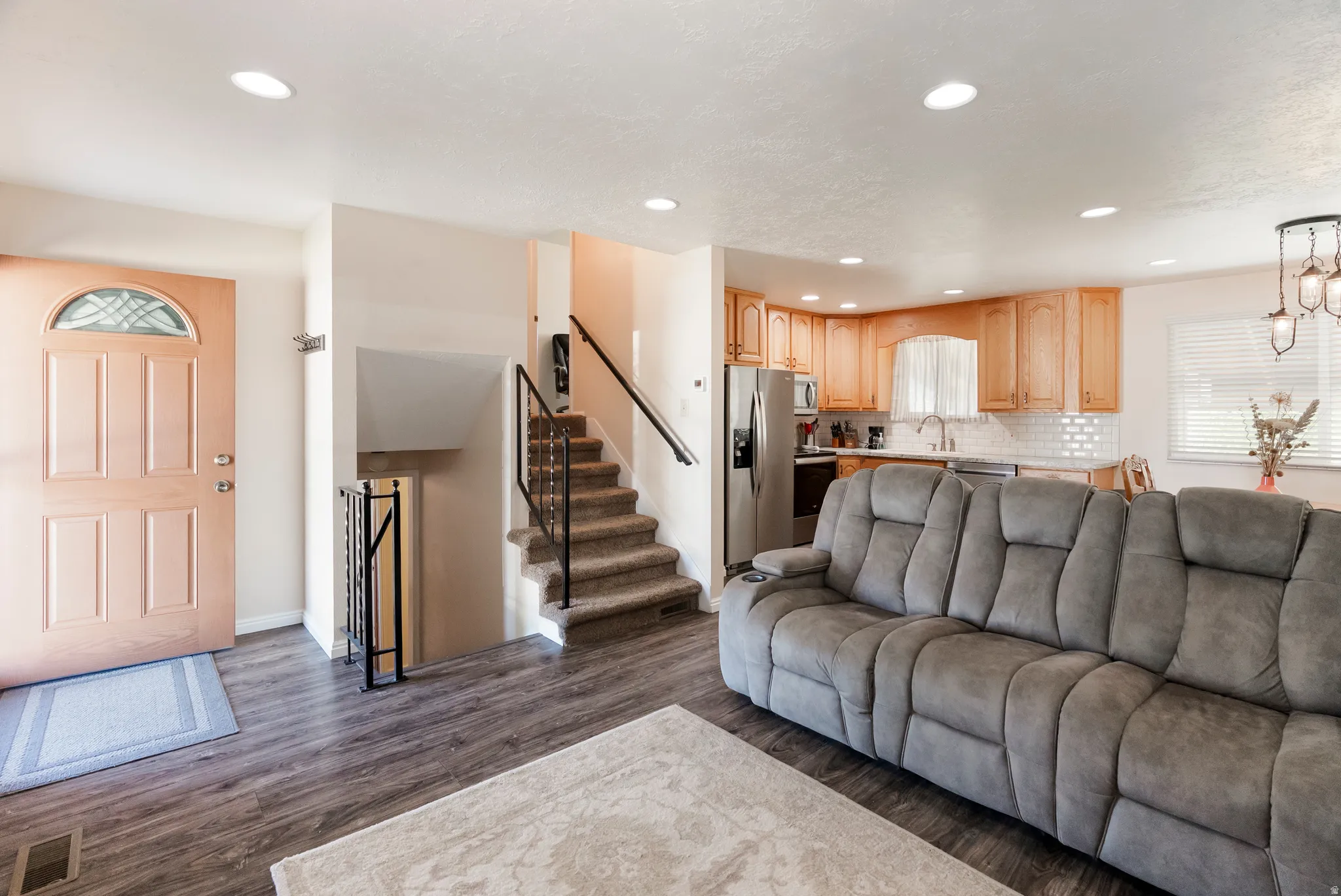 Open-concept living area featuring wood-finish flooring, recessed lighting, and a kitchen with light wood cabinetry, stainless steel appliances, and a subway tile backsplash