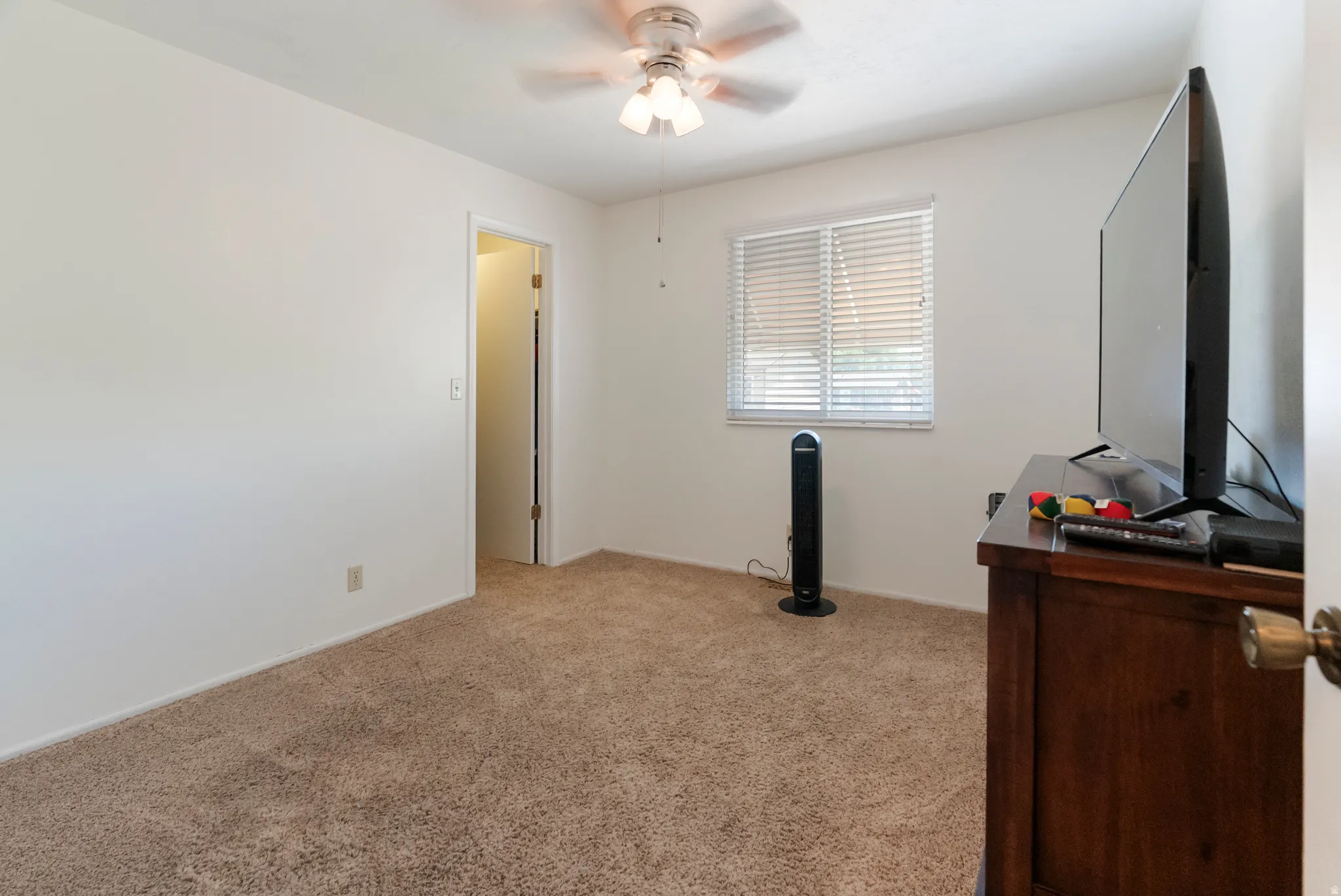 Neutral tone wall color, plush carpet flooring, natural light through the window, and a ceiling fan with integrated lighting
