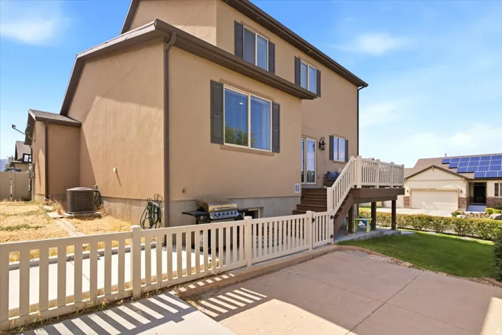 Rear exterior featuring a two-story stucco facade, elevated deck with white railing, paved patio area, and a white picket fence