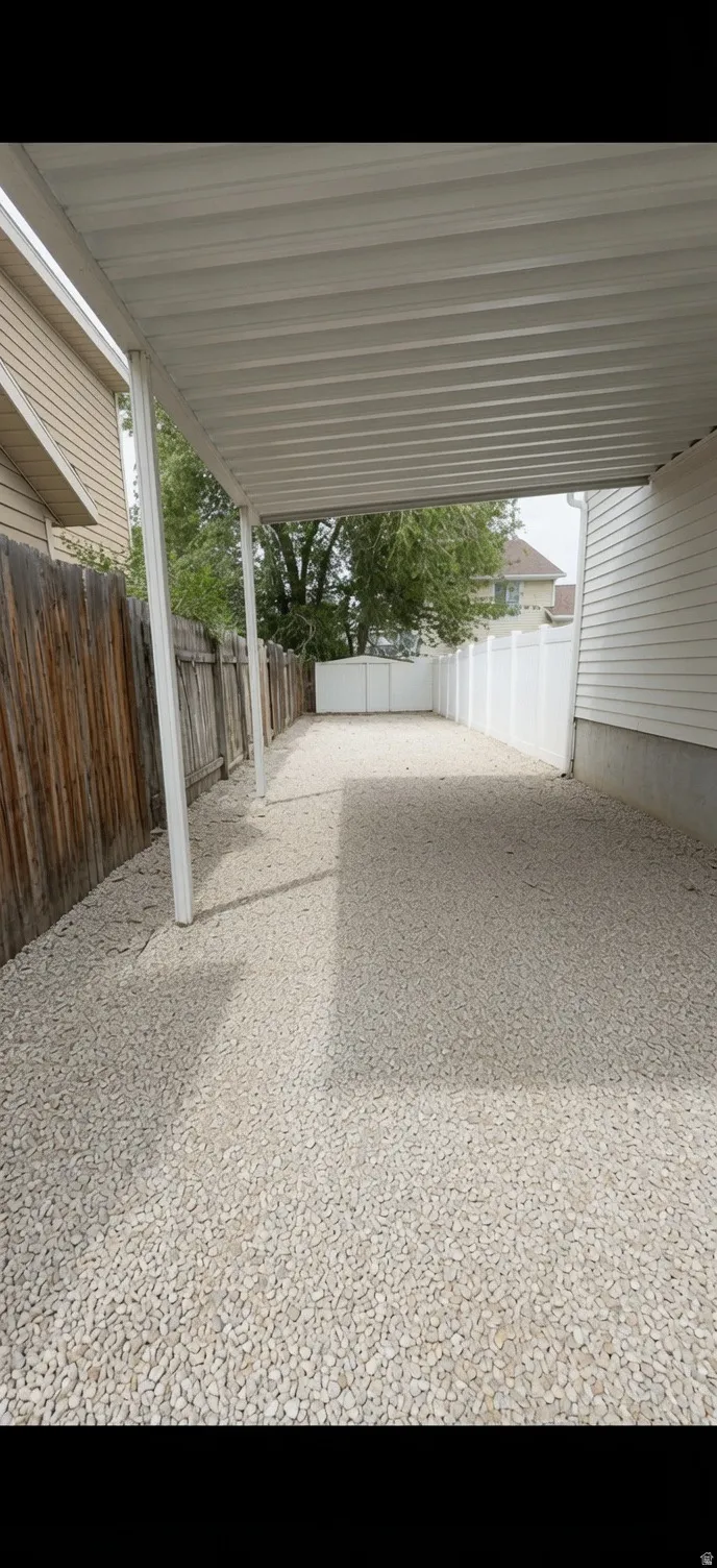 Covered gravel parking area featuring a white metal awning, support posts, and perimeter fencing