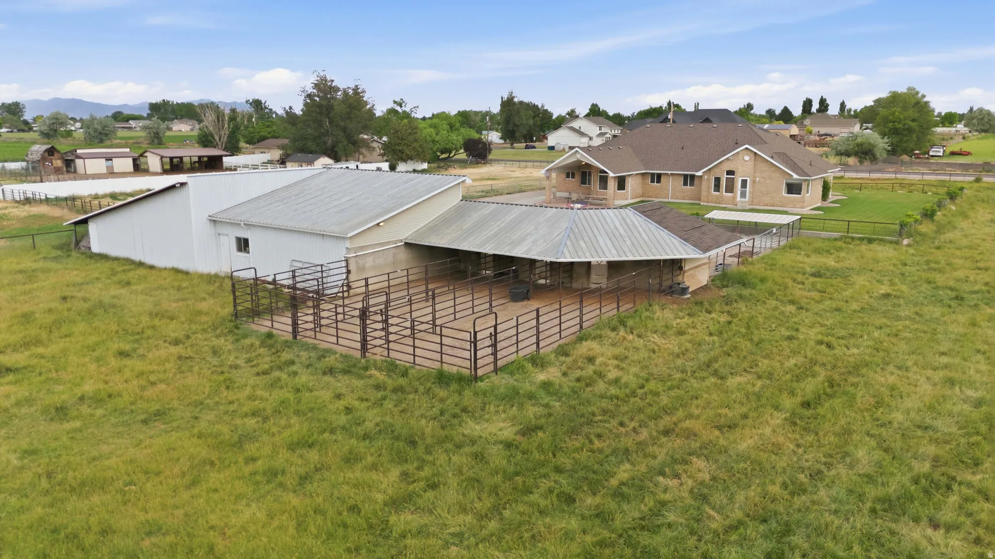 Expansive property featuring a brick exterior residence with a brown shingle roof, accompanied by a large white metal barn with a pitched roof, and a covered animal enclosure with metal fencing