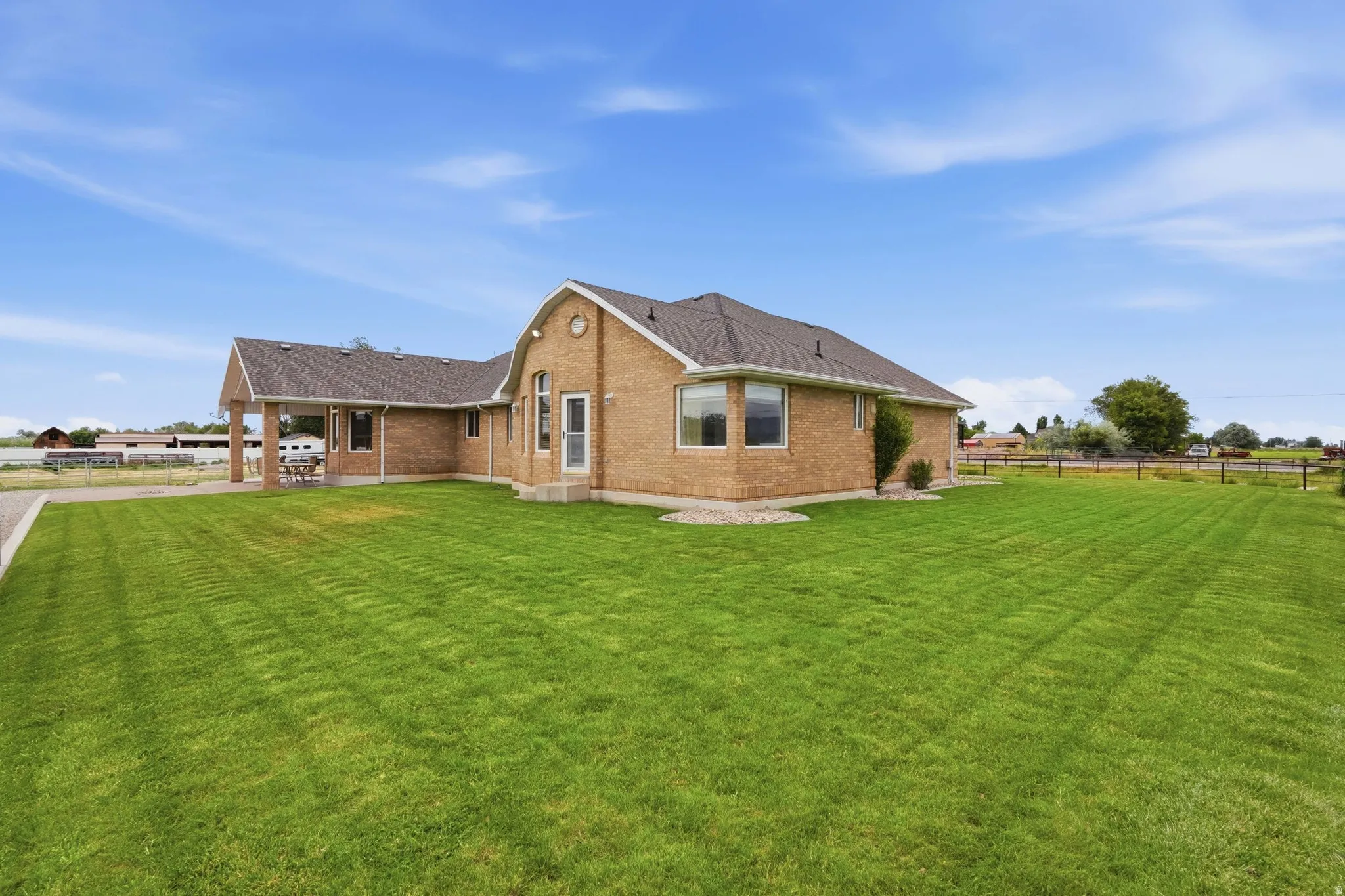 Expansive green lawn surrounds a brick residence featuring multiple windows, a covered patio area, and a paved driveway