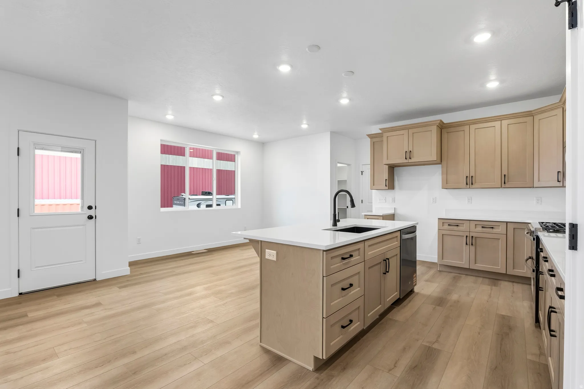 Open-concept kitchen featuring an island with an undermount sink, light wood-finish cabinetry with black hardware, white countertops, and wood-finish flooring