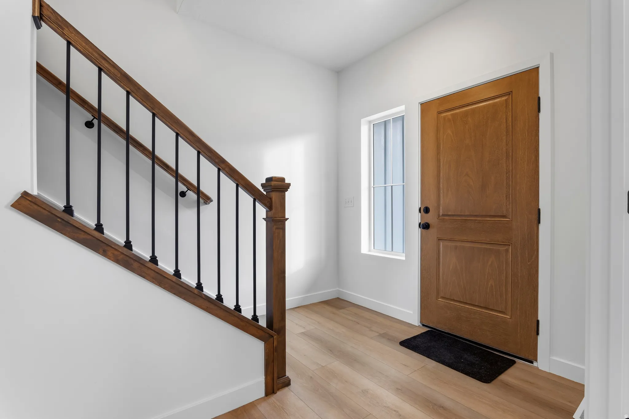 Entryway featuring a wood-panel door with matte black hardware, wood-finish flooring, a side window with grid detailing, and a staircase with wood handrails and black metal spindles