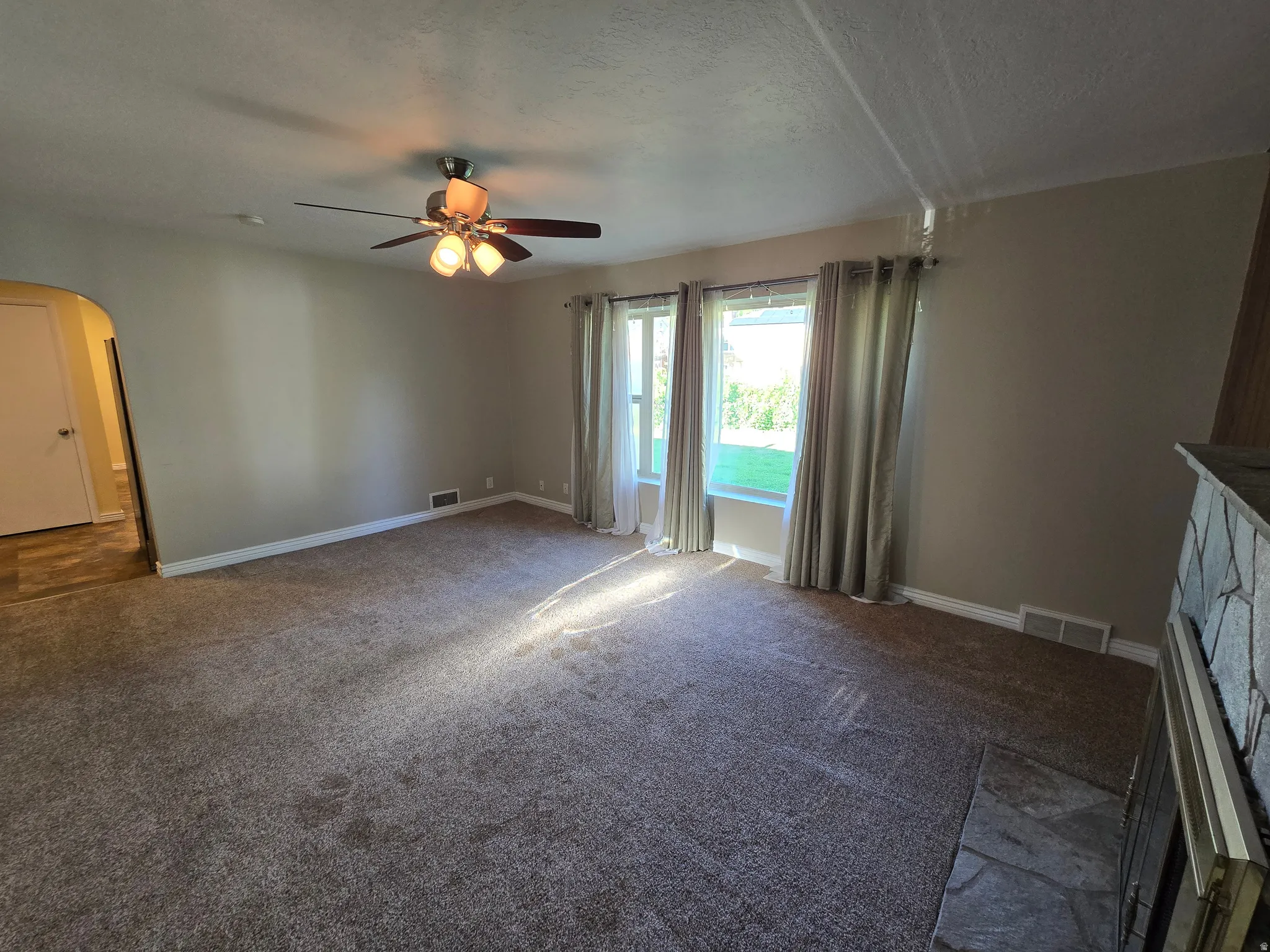 Spacious carpeted room featuring a ceiling fan with integrated lighting, multiple windows with drapes, an archway entrance, and a stone-clad fireplace