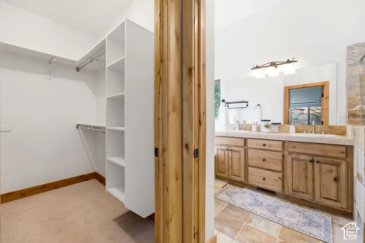 Walk-in closet with built-in shelving, carpet flooring, and wood trim, alongside a bathroom featuring a double vanity with wood cabinetry, light-toned countertop, and large mirror