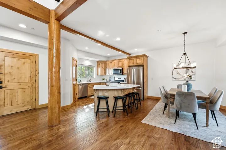 Open-concept living area featuring exposed wood beams, wood-finish flooring, a kitchen with stainless steel appliances, light-toned cabinetry, and a central island with bar seating